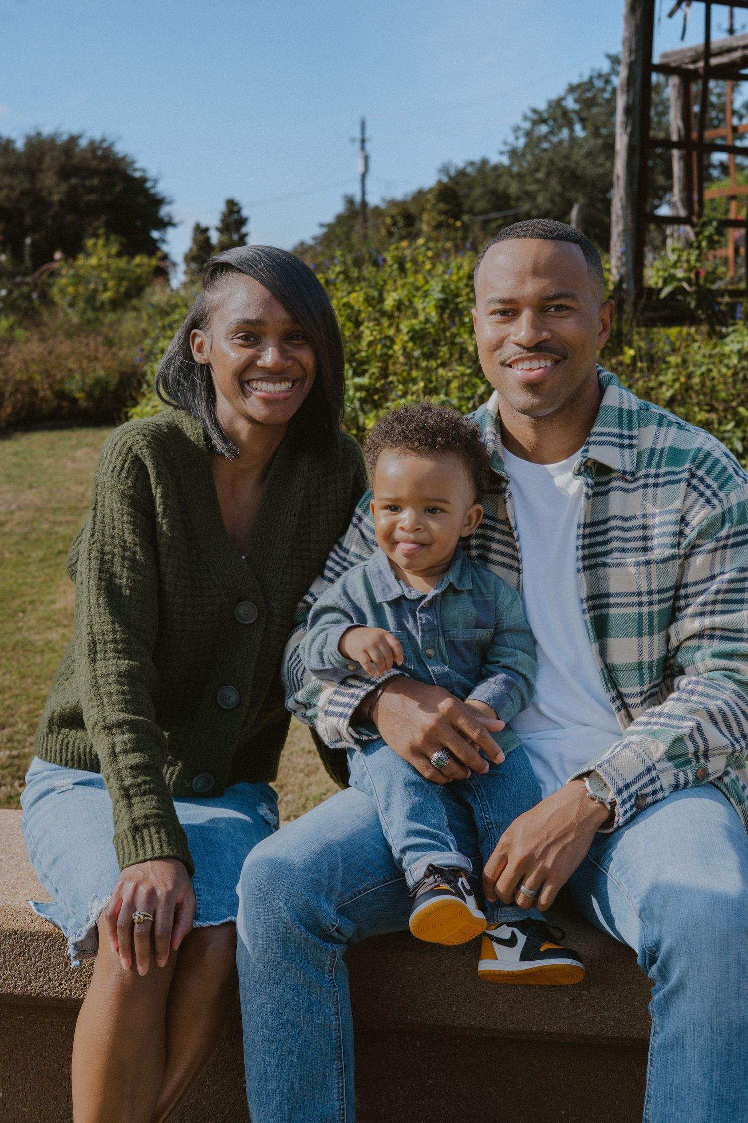 Happy Black family of three sits together outdoors, parents smiling with baby boy on dad's lap.