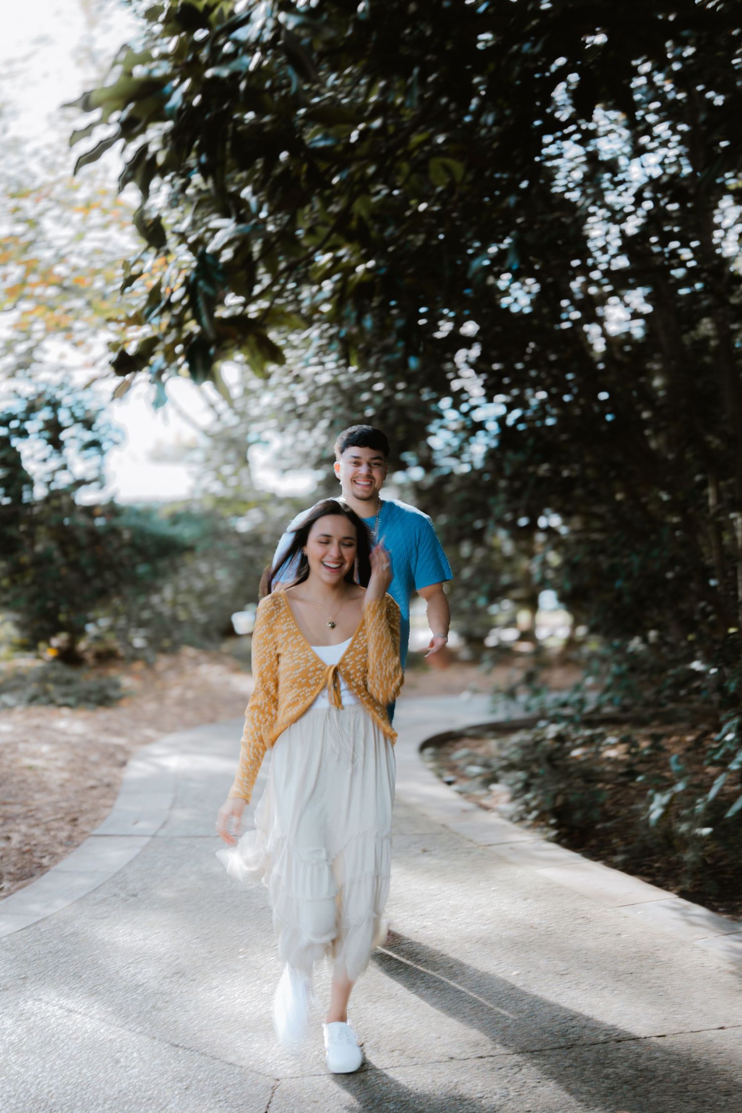 A couple walking on a paved path surrounded by trees, with one wearing a yellow cardigan and white dress.