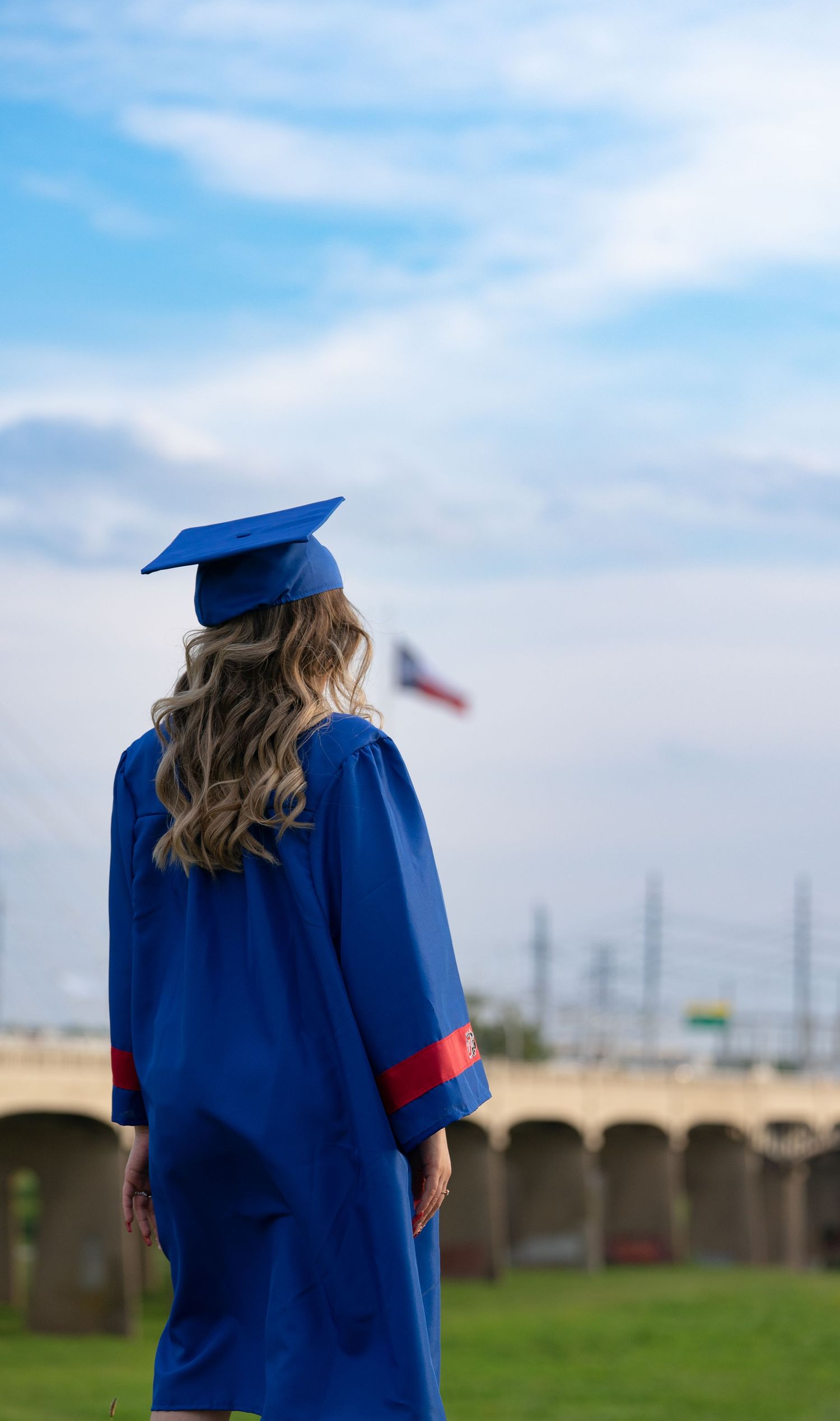 Graduate in blue cap and gown stands facing away from camera in outdoor setting with cloudy sky background.