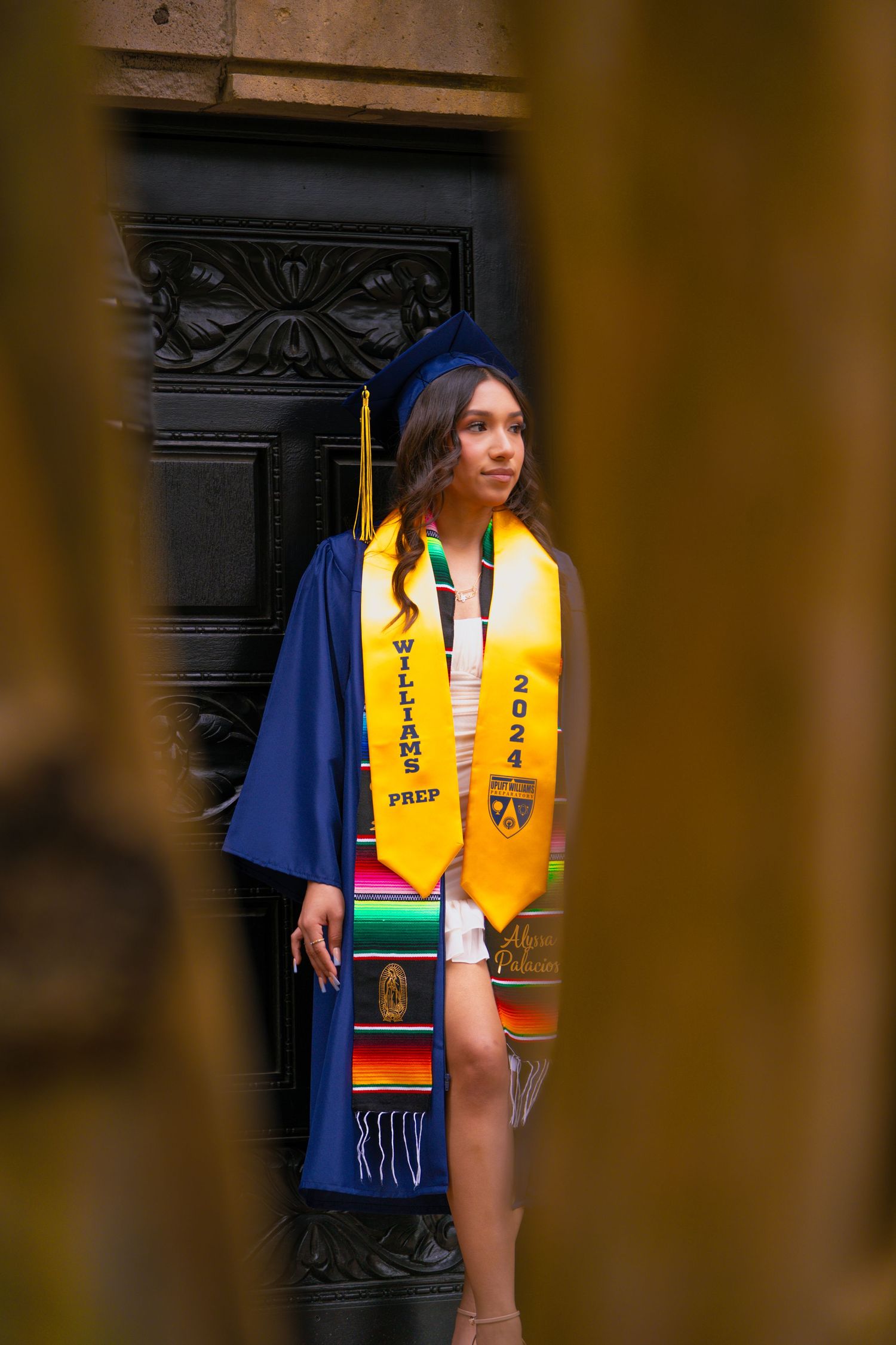 Graduate wearing blue gown and yellow stole poses against dark doorway in artistic graduation photo.