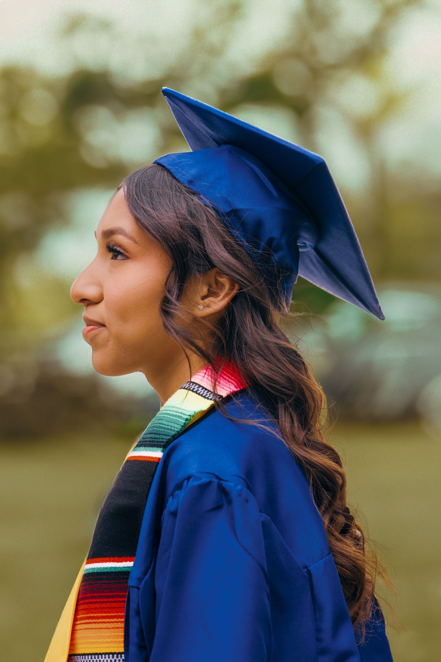 Graduate in blue cap and gown with colorful serape graduation stole smiles while looking to the side outdoors.