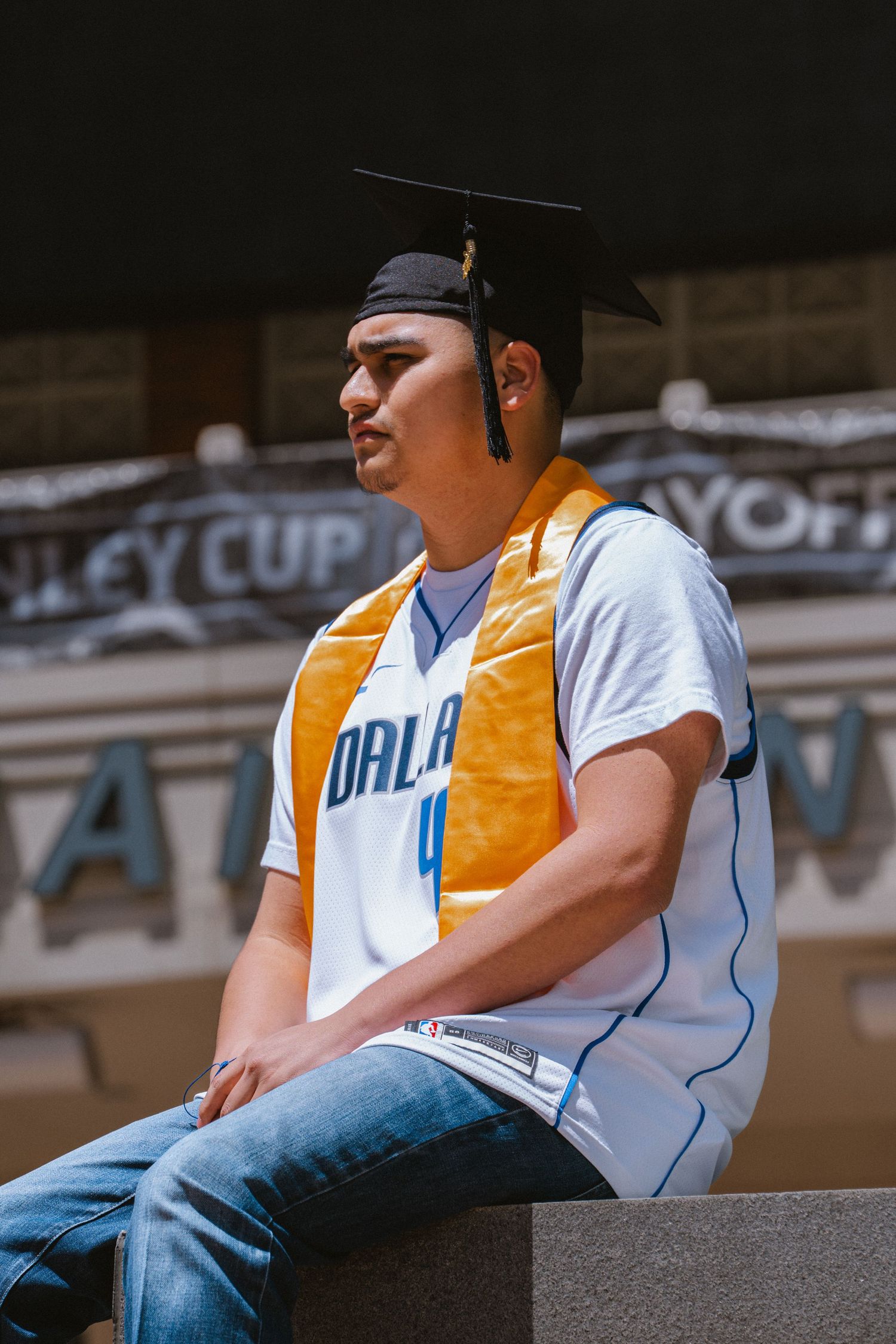 Young man in graduation cap and gold sash wearing Orlando Magic jersey sits outdoors at Stanley Cup Playoffs venue.