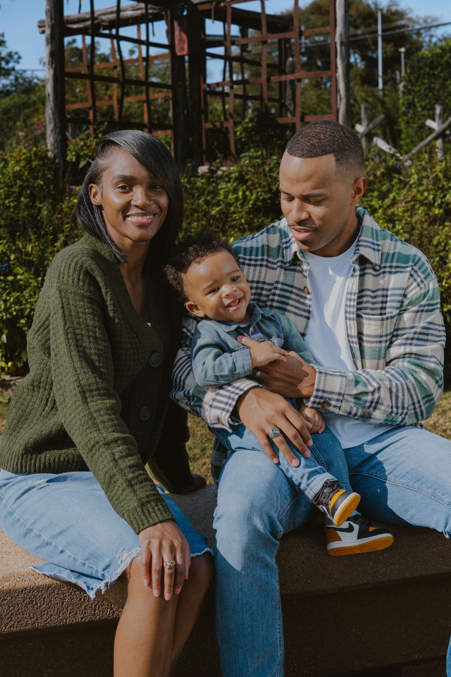 A happy family of three sits outdoors, with parents smiling and holding their laughing toddler on a sunny day.