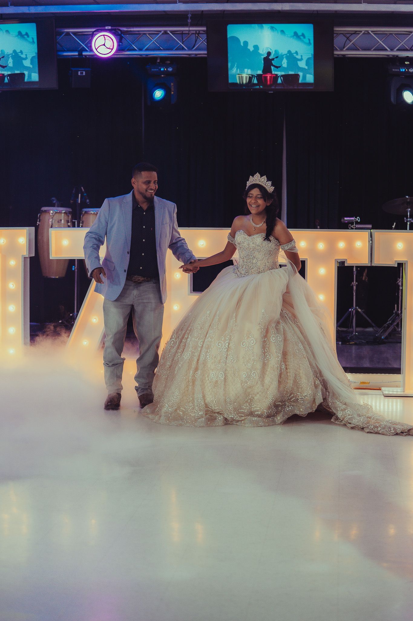 A couple shares their first dance at a quinceañera, the young woman wearing a stunning ballgown and crown on a foggy dance floor.