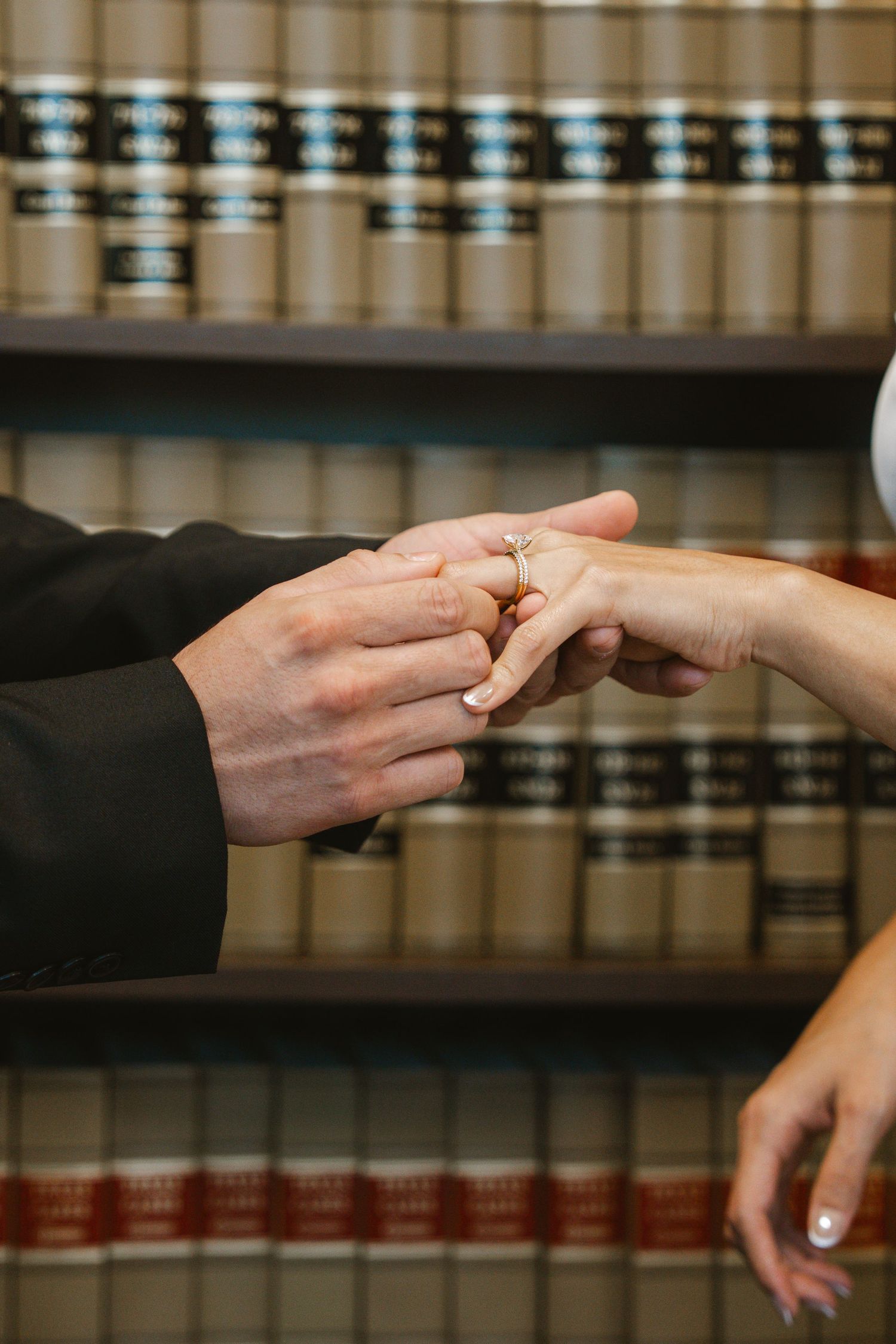 A groom placing a gold wedding ring on bride's finger in front of law library bookshelves.