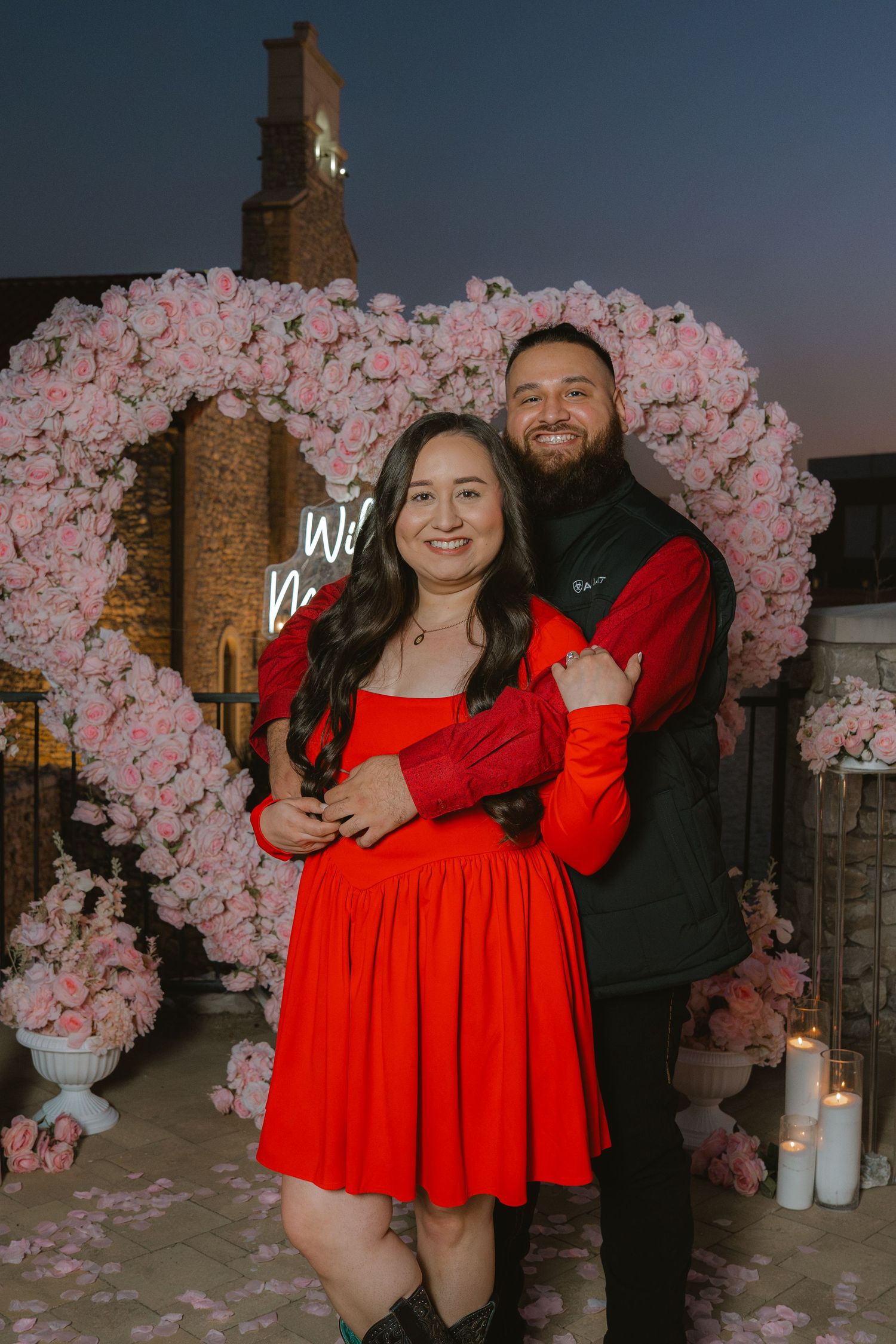 Couple in matching red outfits embracing before a pink rose heart arch with candles at a romantic proposal setup.