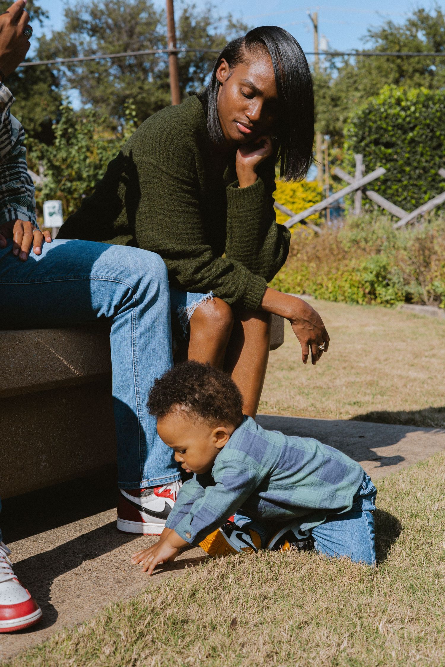 A mother in a green sweater watches her toddler crawl on grass in a sunny outdoor park setting.