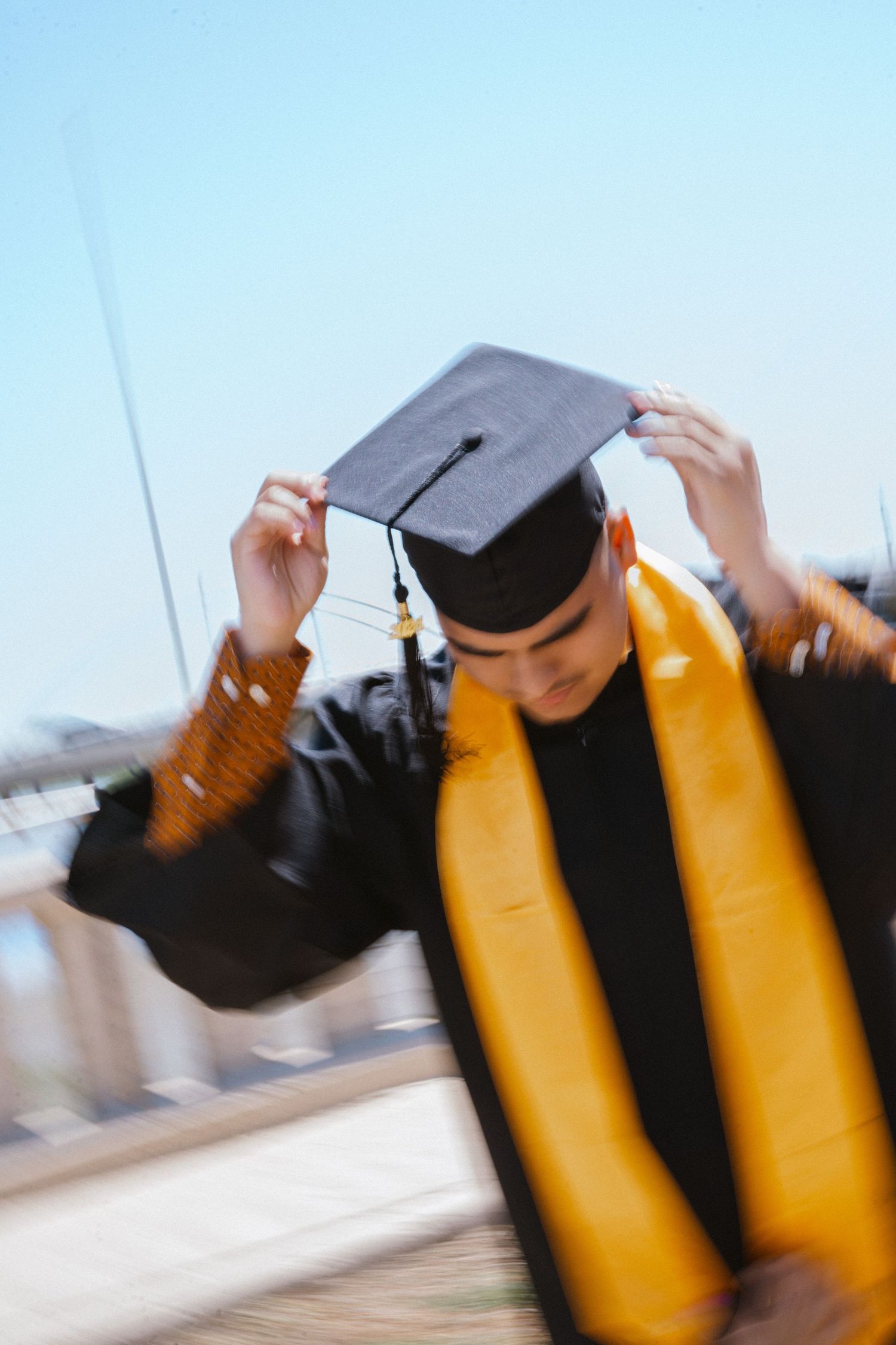 Graduate in black cap and gown with gold stole adjusting mortarboard, motion blur effect outdoors.