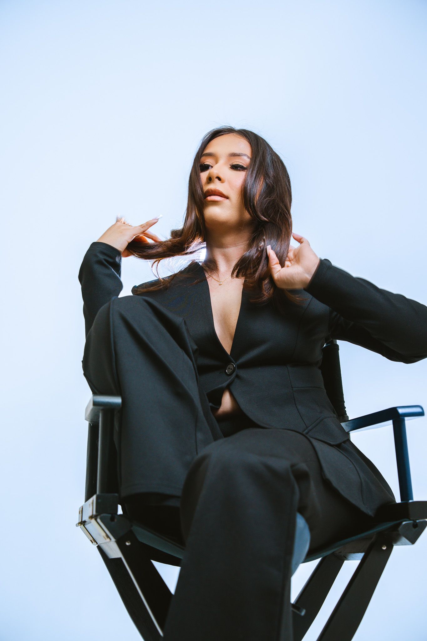 Model in black suit poses dramatically on director's chair against light blue background.
