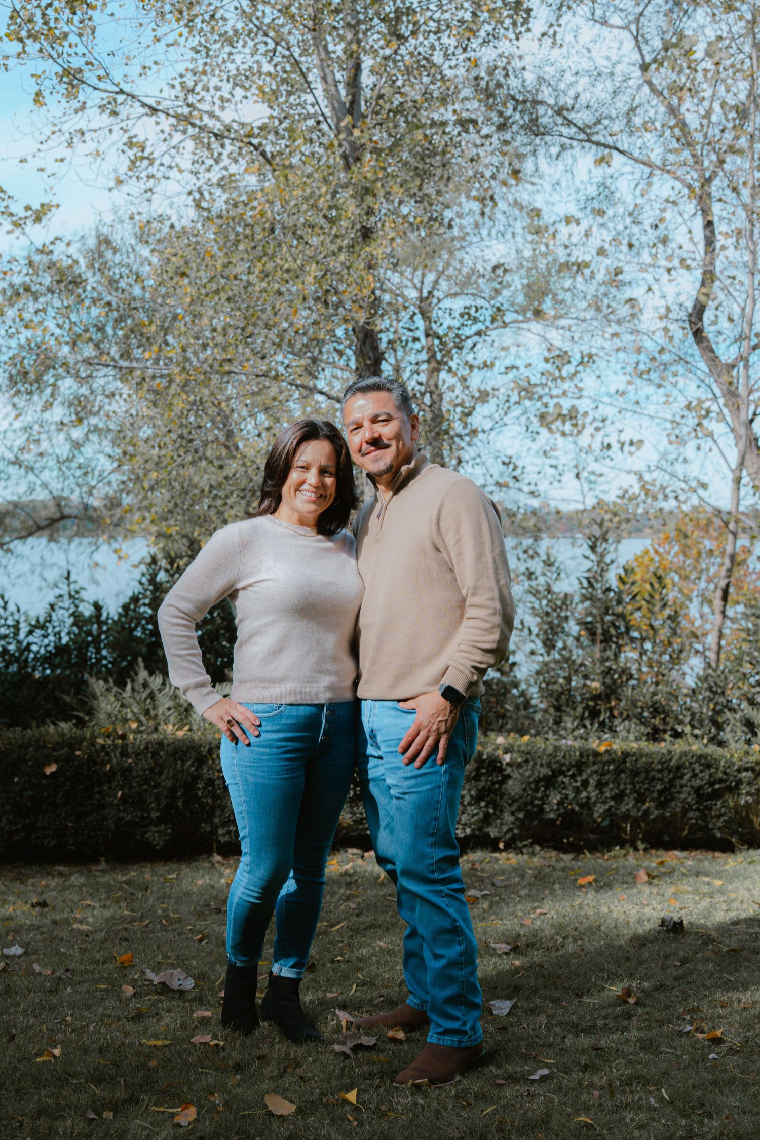 A couple wearing matching beige sweaters and blue jeans pose playfully together in an outdoor autumn setting.