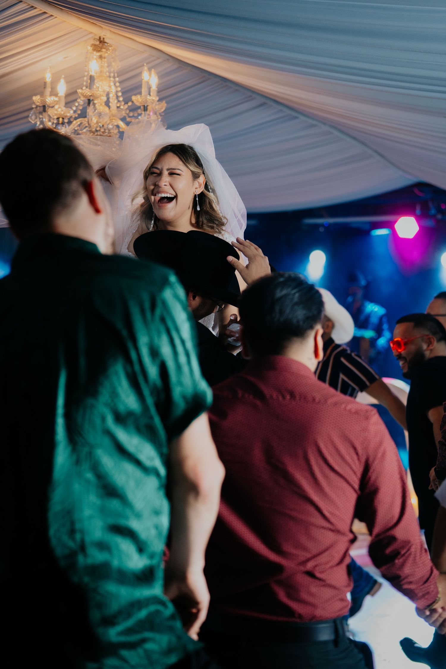 People dancing and celebrating at a lively party under a tent with colorful lighting and a chandelier.