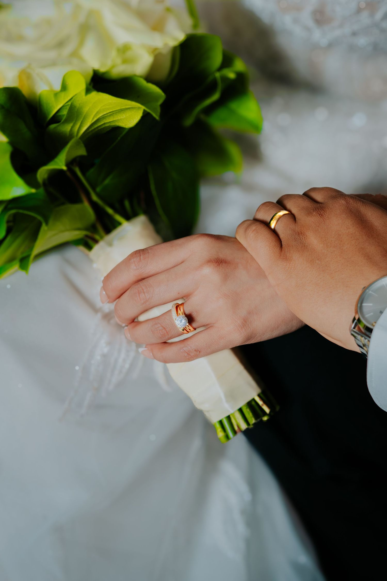 Close-up shot of hands holding a wedding bouquet with green foliage and white flowers against a light background.