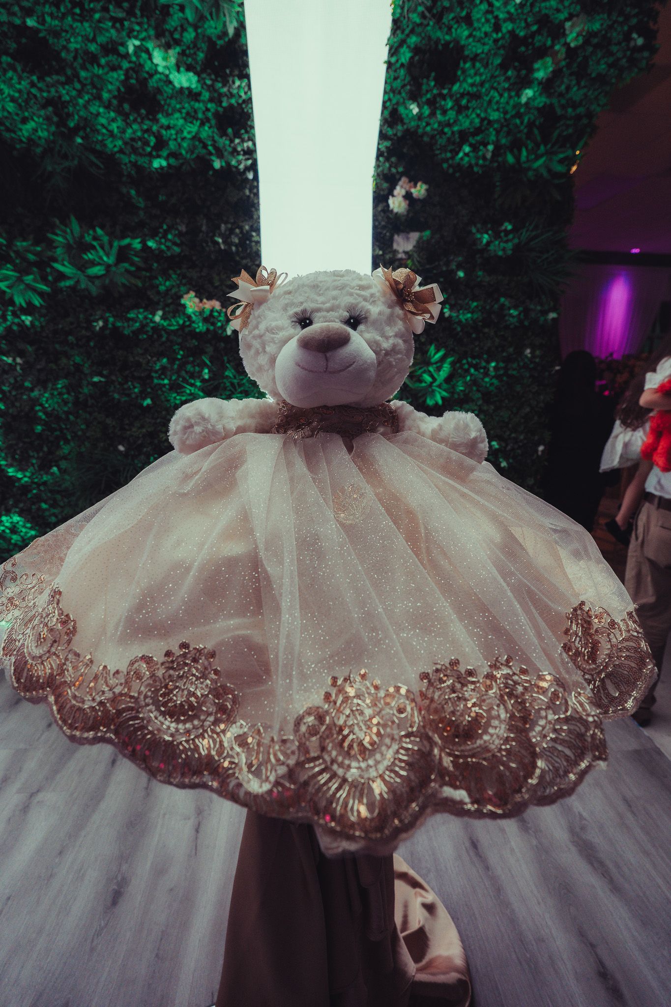 A white teddy bear wearing an elegant gold-trimmed tulle ballgown dress displayed at a decorated event venue.