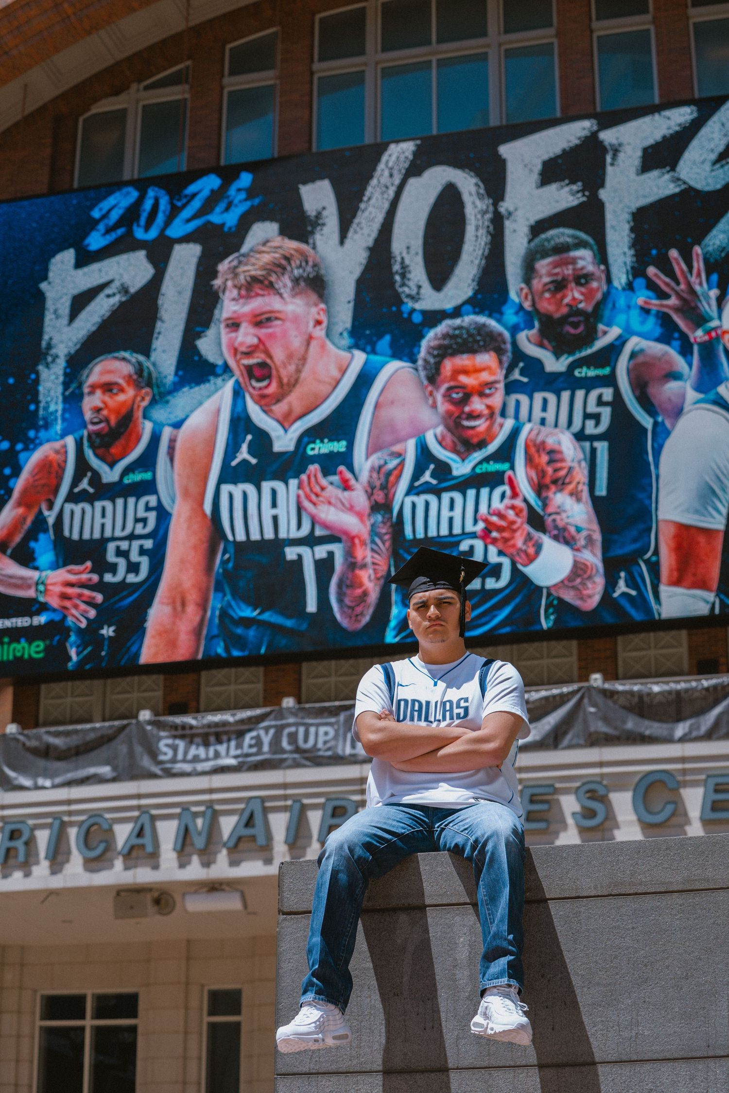Dallas Mavericks fan poses arms crossed outside arena beneath 2024 NBA Playoffs promotional billboard.