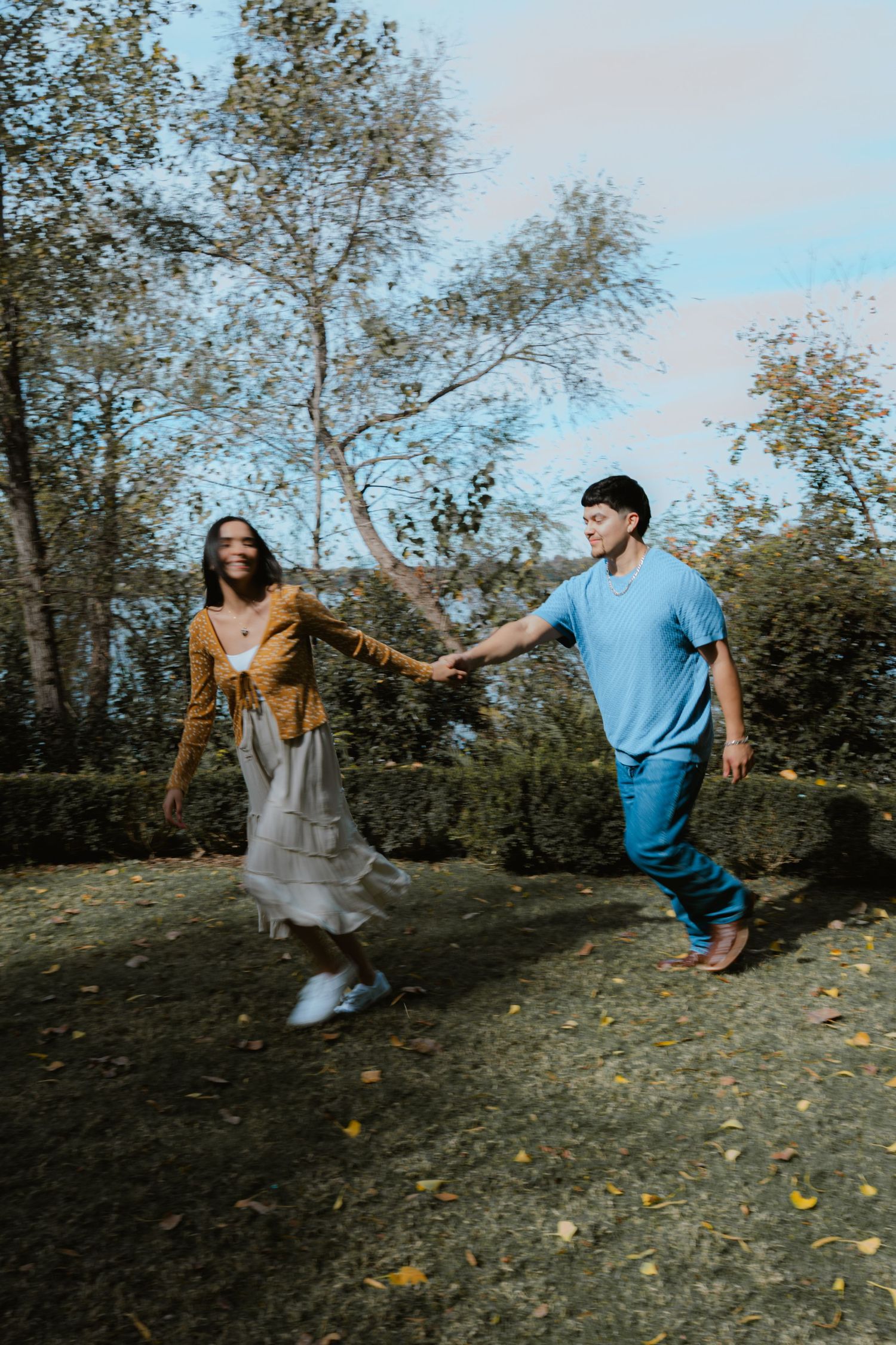 A couple dances playfully in a grassy outdoor setting with autumn leaves scattered on the ground.