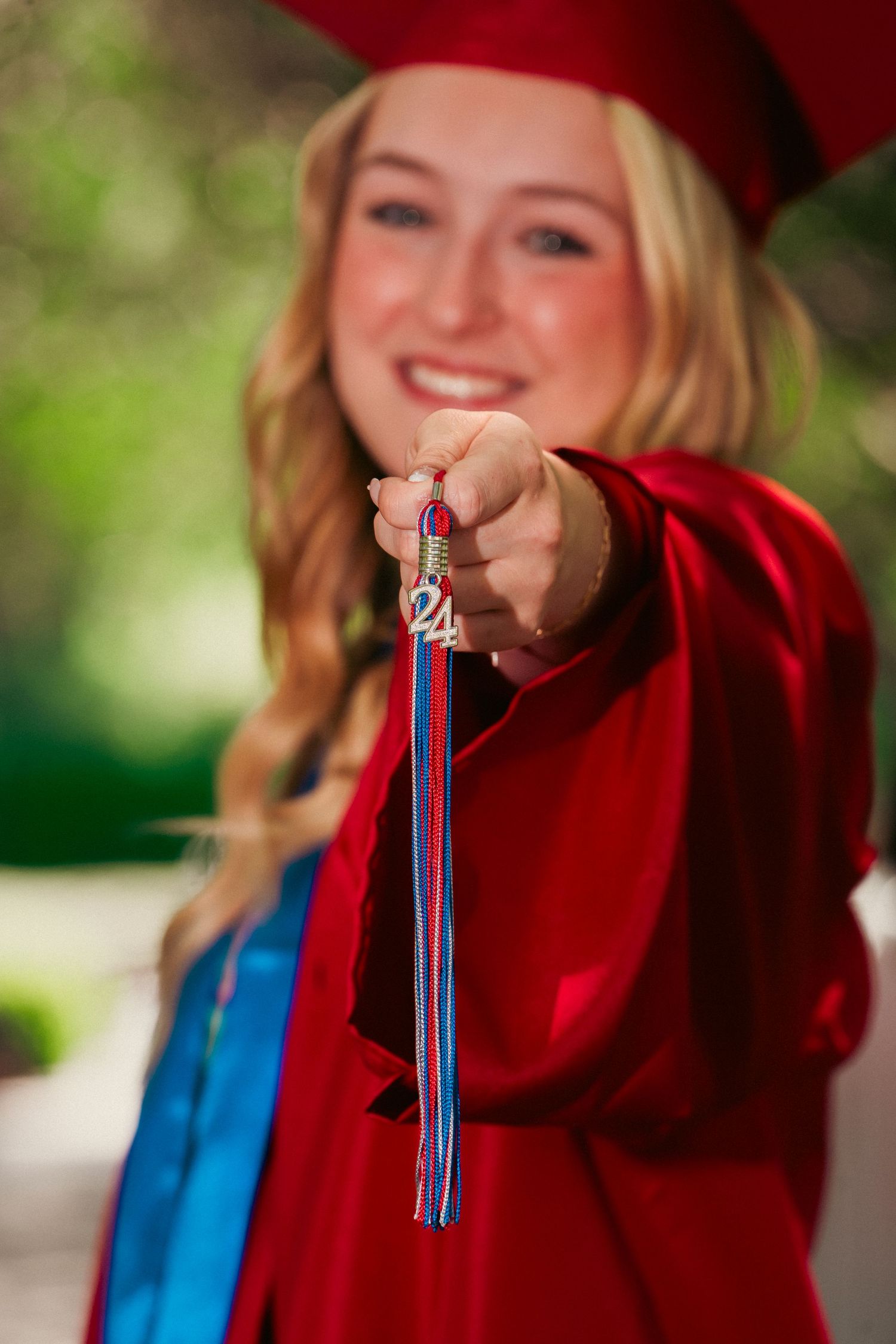 Graduate in red gown holds up a graduation tassel with blue and red colors against a blurred outdoor background.