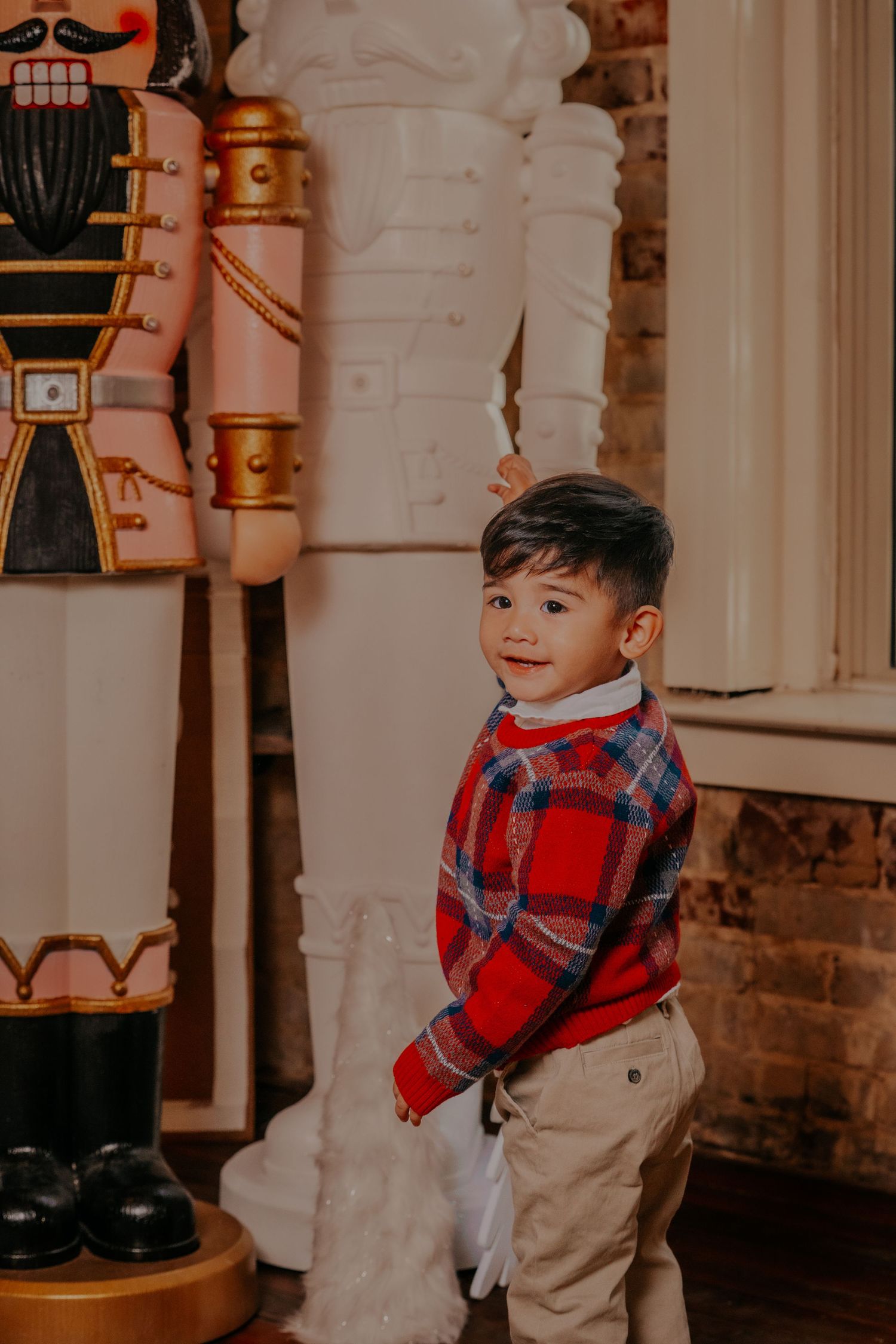 Young child in plaid scarf and khaki pants stands in front of rustic brick wall with holiday decor.