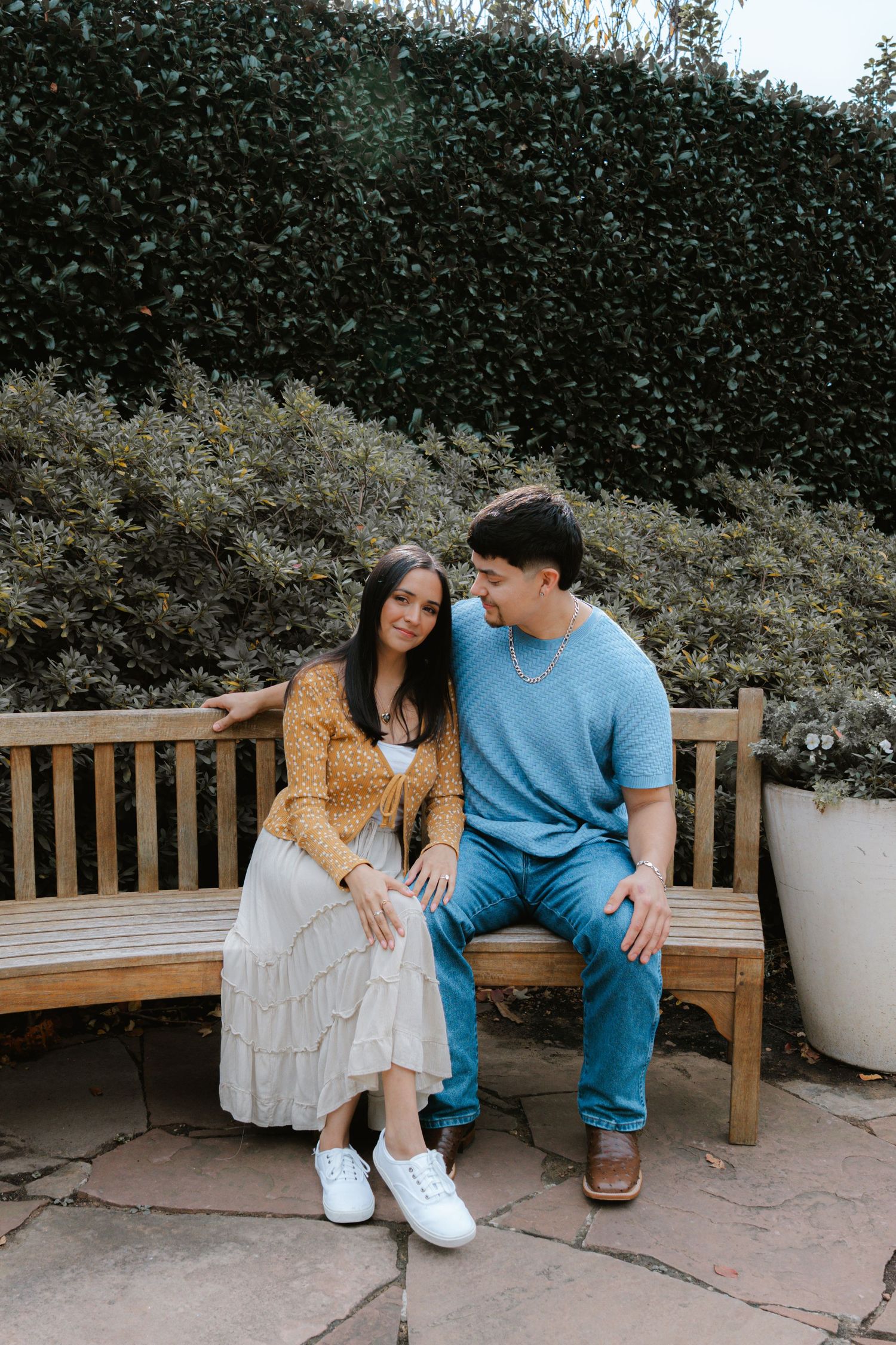 A couple sits together on a wooden bench in a garden setting, wearing casual outfits of blue and yellow.