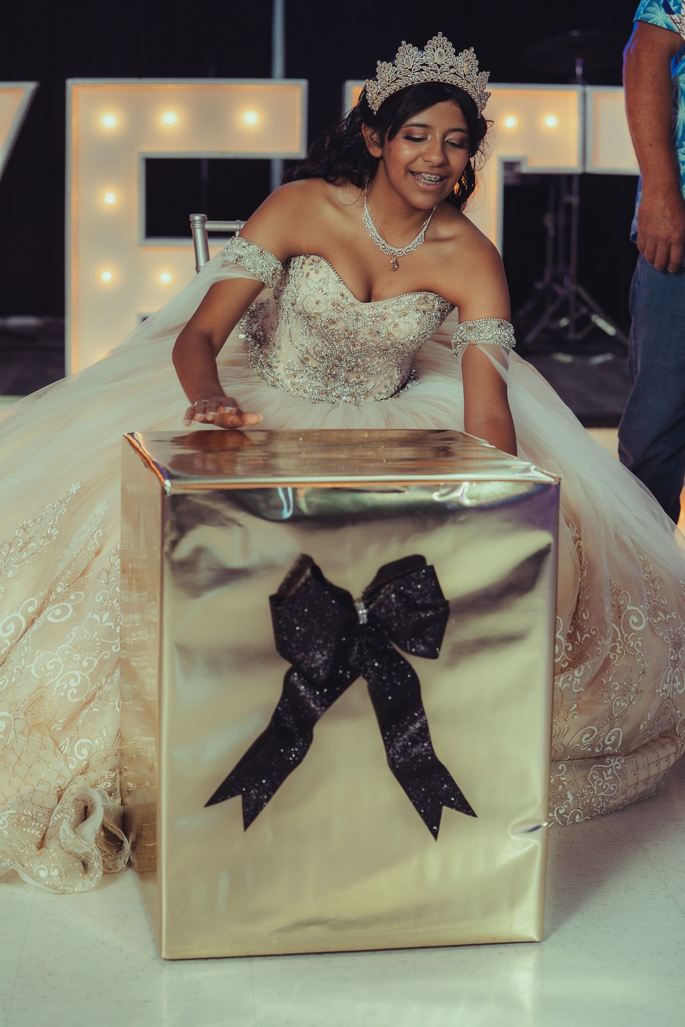 A quinceañera girl in a white lace gown and tiara smiles beside a large gold gift box with a black bow.