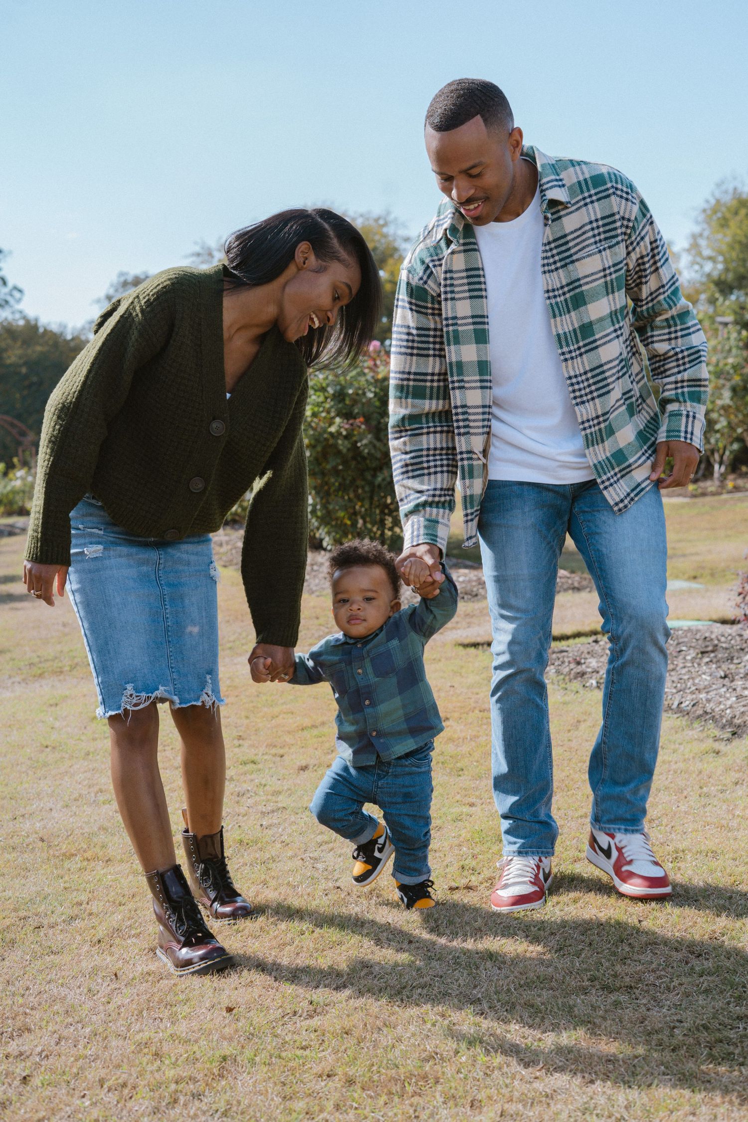 A joyful Black family holds their toddler's hands as he takes steps on a sunny green lawn.