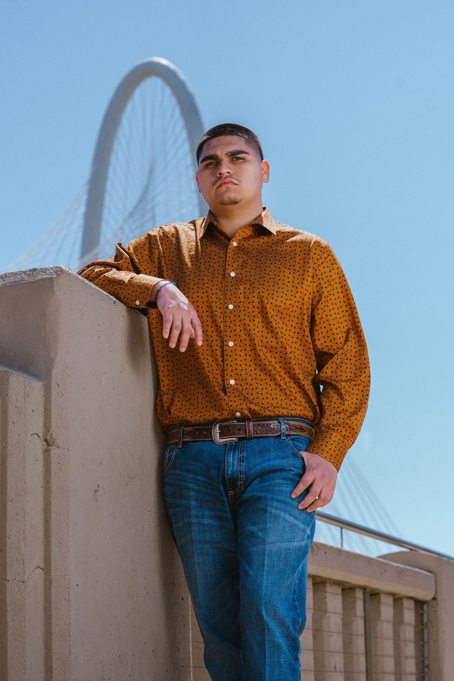 Young man in orange patterned shirt and jeans leans against concrete wall with cable bridge behind him.