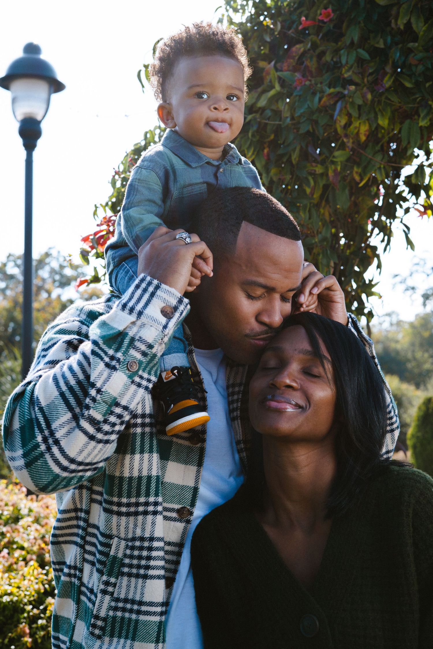 Happy family with father kissing mother while toddler sits on his shoulders in a garden park.