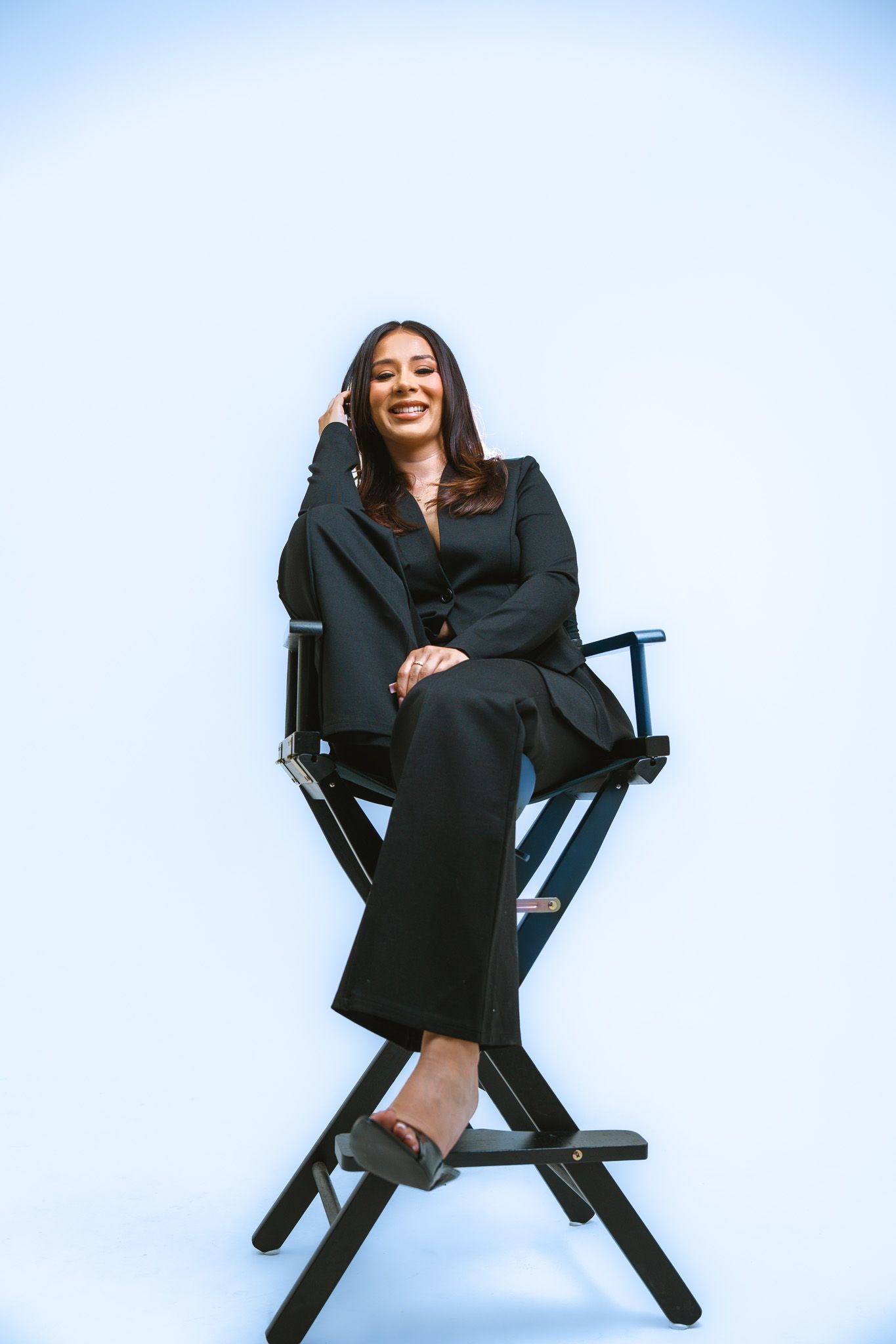 Professional studio portrait of someone in a black business suit sitting casually in a director's chair against white backdrop.