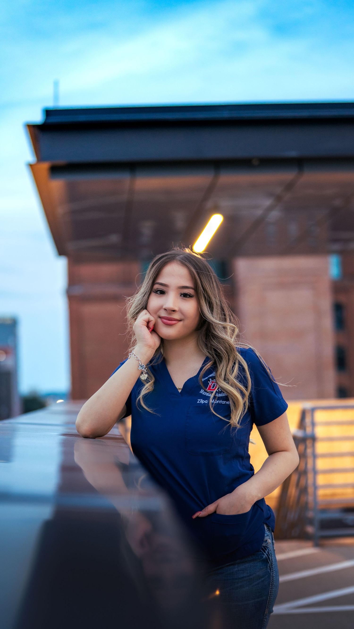 A person in navy blue attire poses outdoors at dusk with warm lighting in the background.