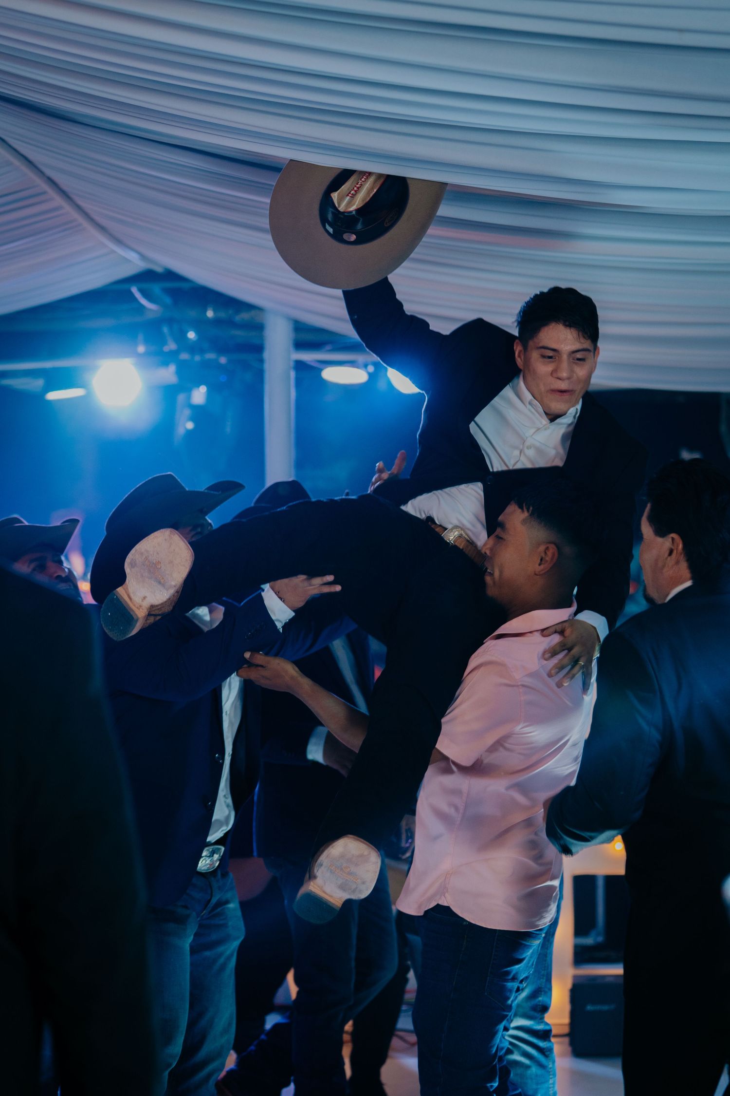 Silhouettes of party guests dancing and mingling under a draped tent ceiling with blue mood lighting.