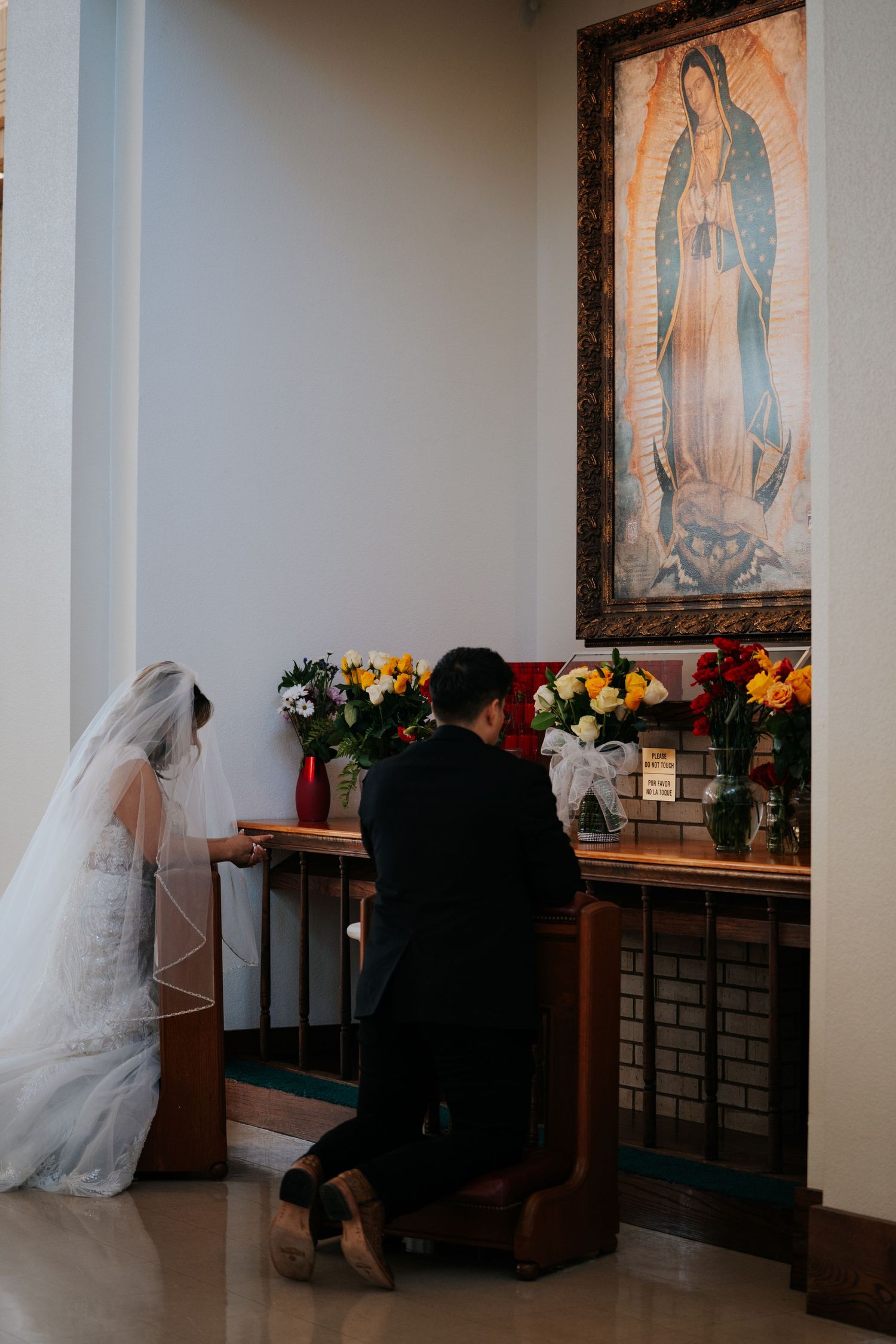 A couple kneels in prayer beneath a religious painting of Our Lady of Guadalupe, surrounded by colorful floral arrangements.