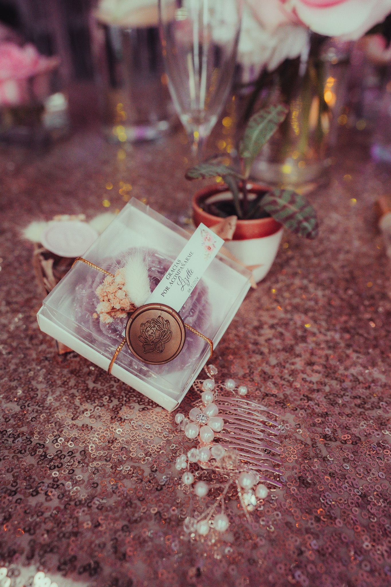 Elegant wedding table detail with gold wax seal gift box on pink sequin tablecloth with florals.
