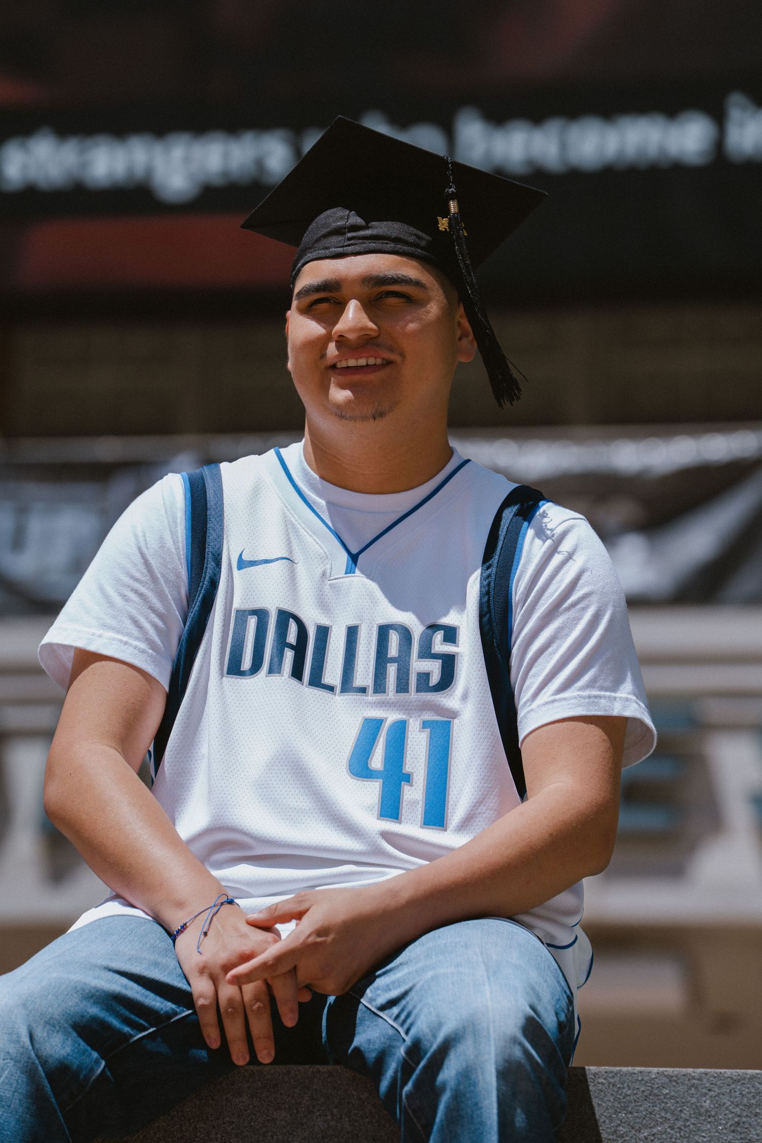 Young man in Dallas Mavericks #41 jersey and graduation cap sits smiling outdoors.