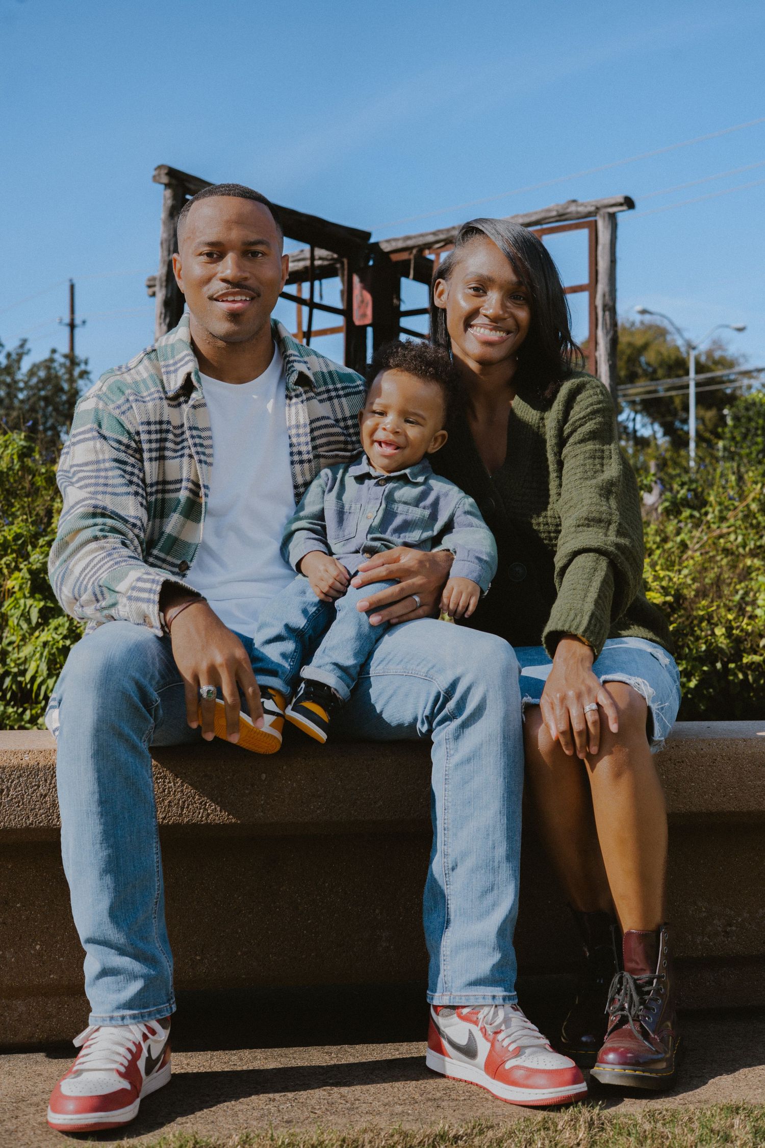 Smiling Black couple sits on an outdoor ledge with their cheerful toddler between them.