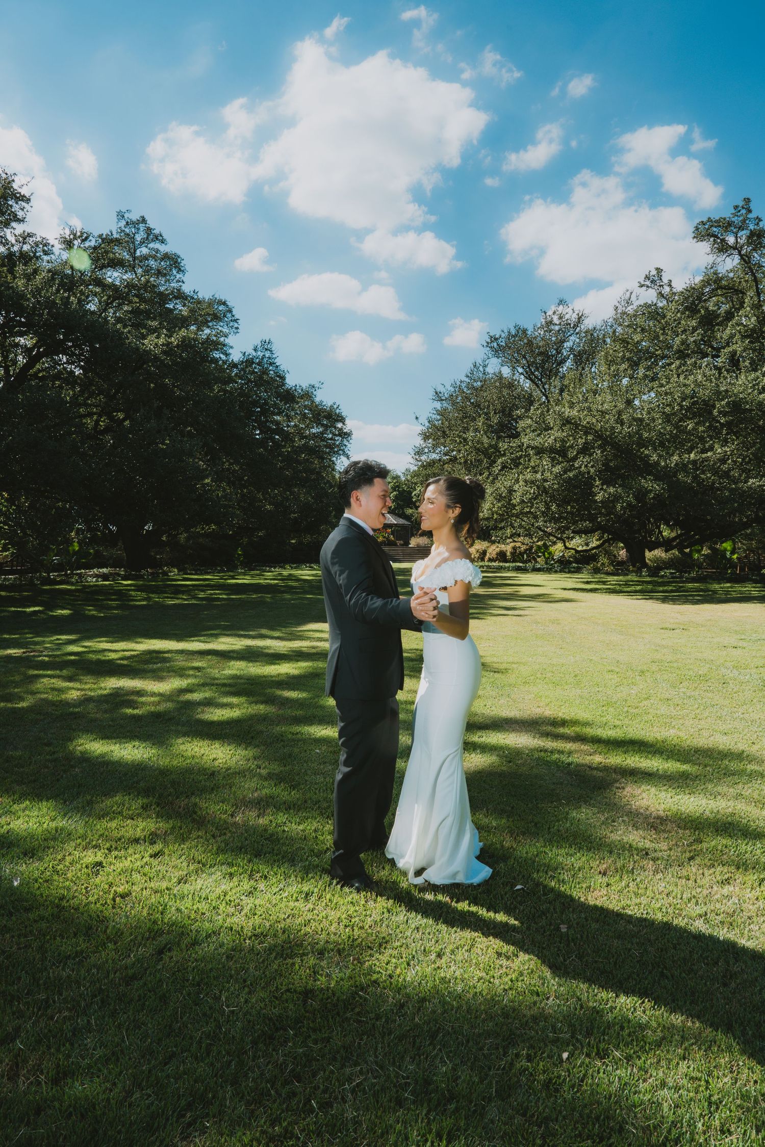 A bride and groom share a romantic moment on a lush green lawn surrounded by trees under a blue sky.