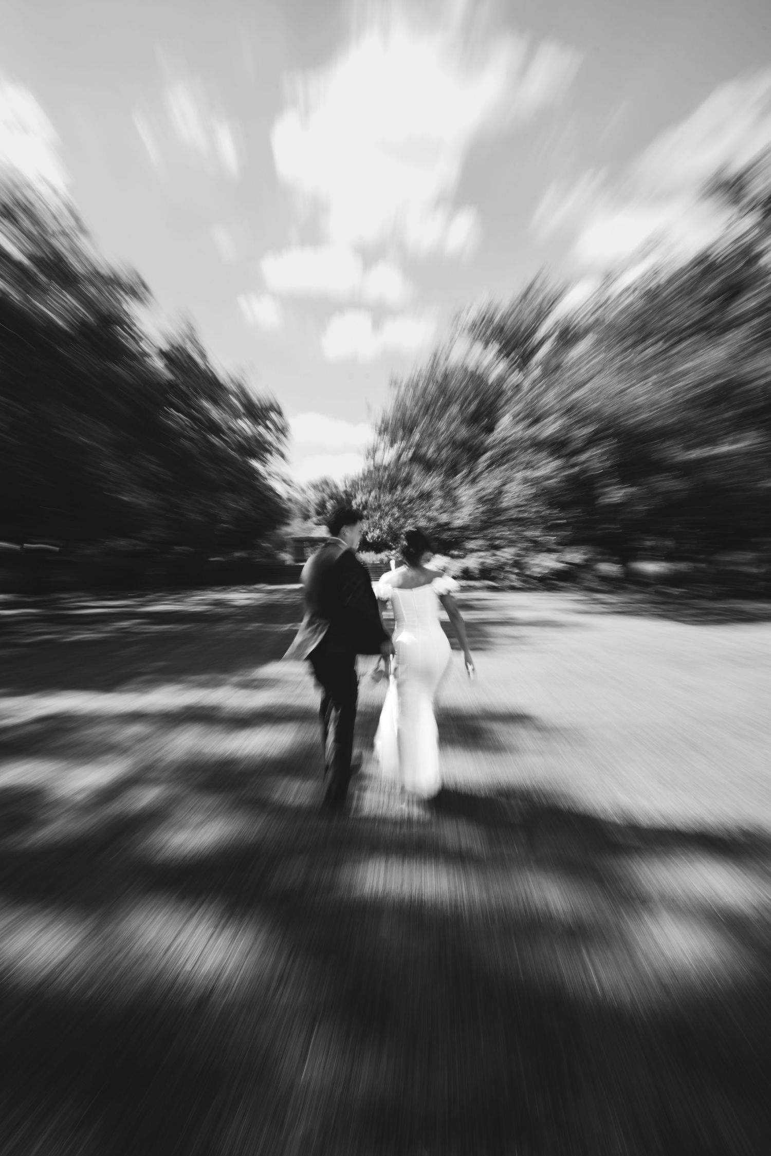 Black and white motion blur photograph of a bride and groom walking through a park pathway creating a dramatic effect.