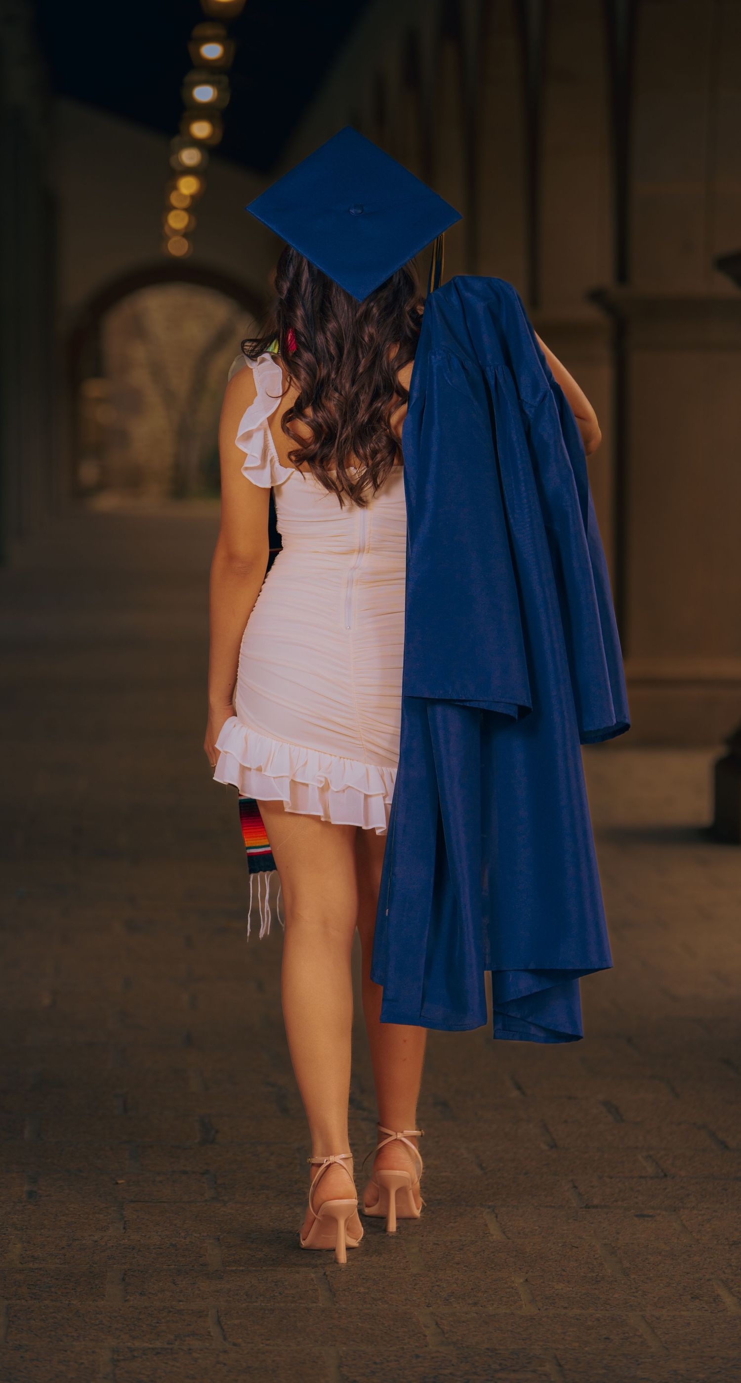 Graduate wearing white dress and blue cap and gown walking on stone pavement in evening light.