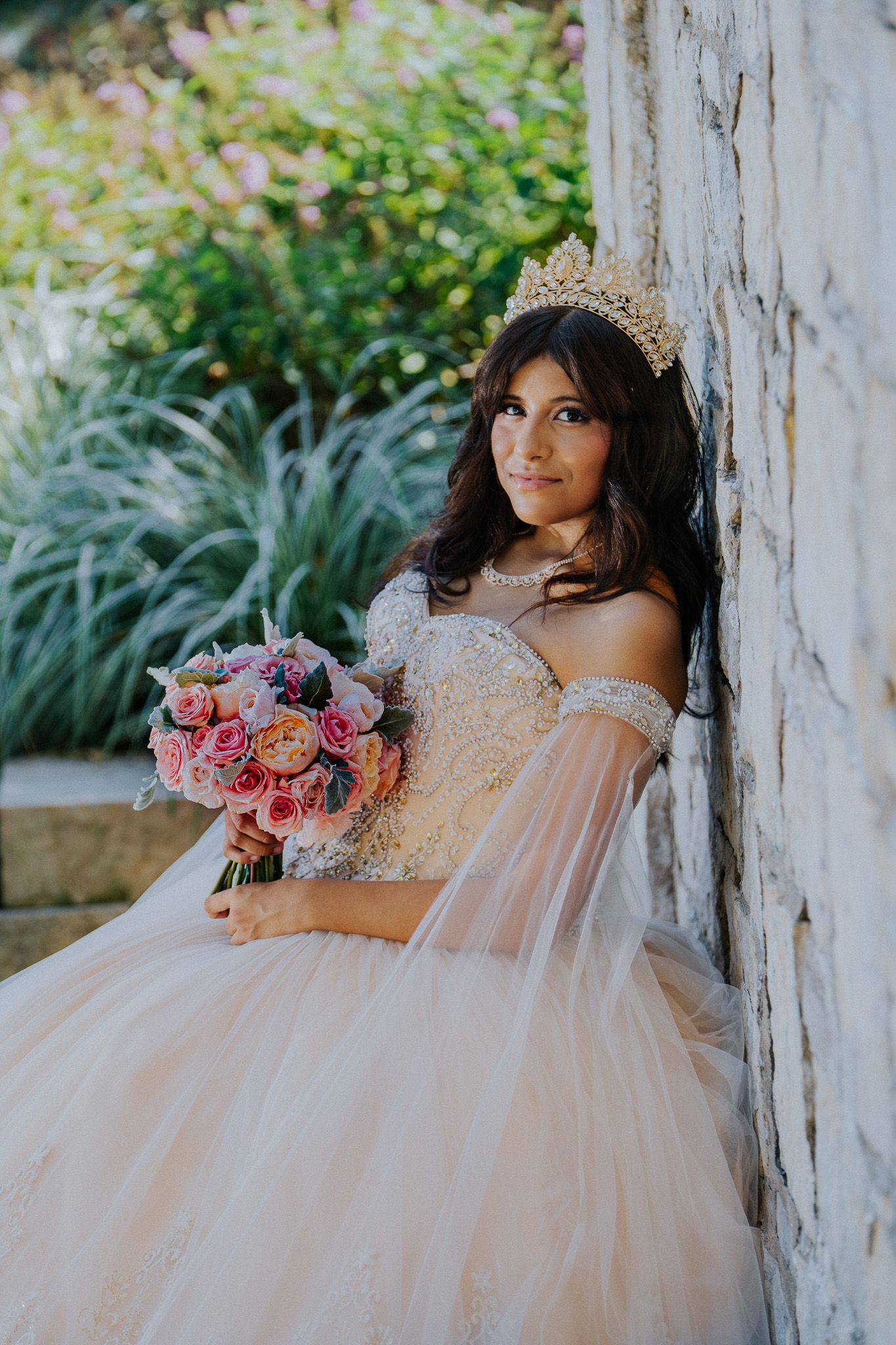 Young woman in a blush ballgown with gold crown holds pink bouquet, leaning against stone wall outdoors.