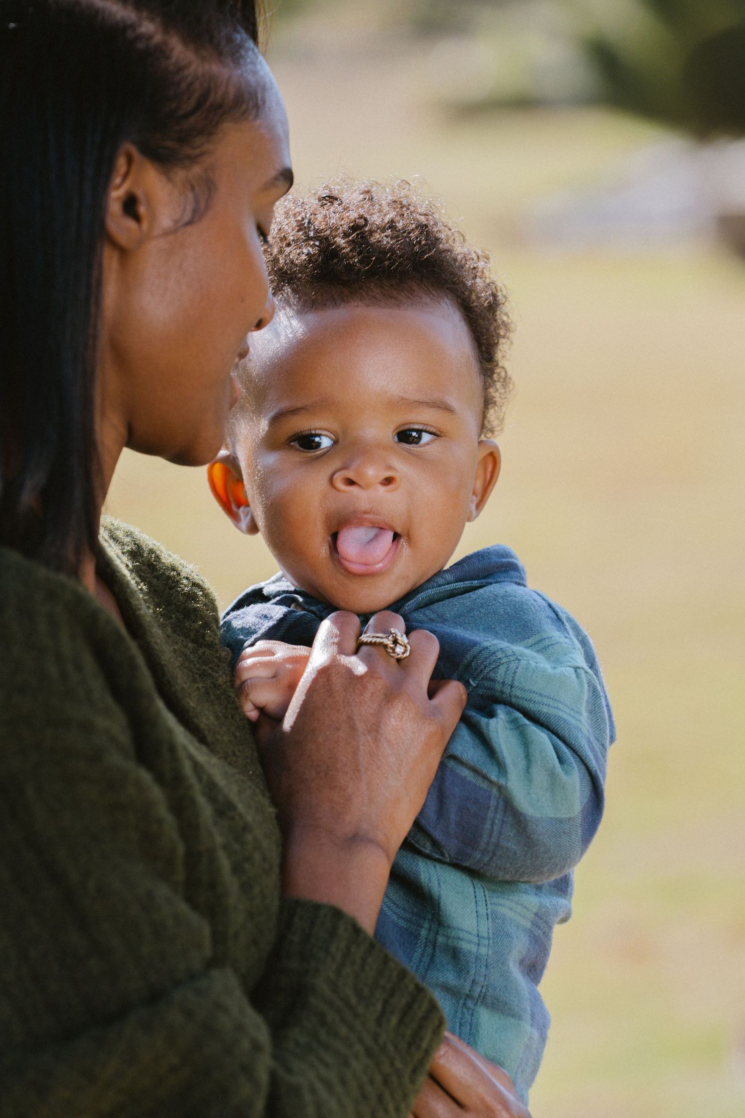 Cute Black baby sticking tongue out while being held by mother in green sweater outdoors.