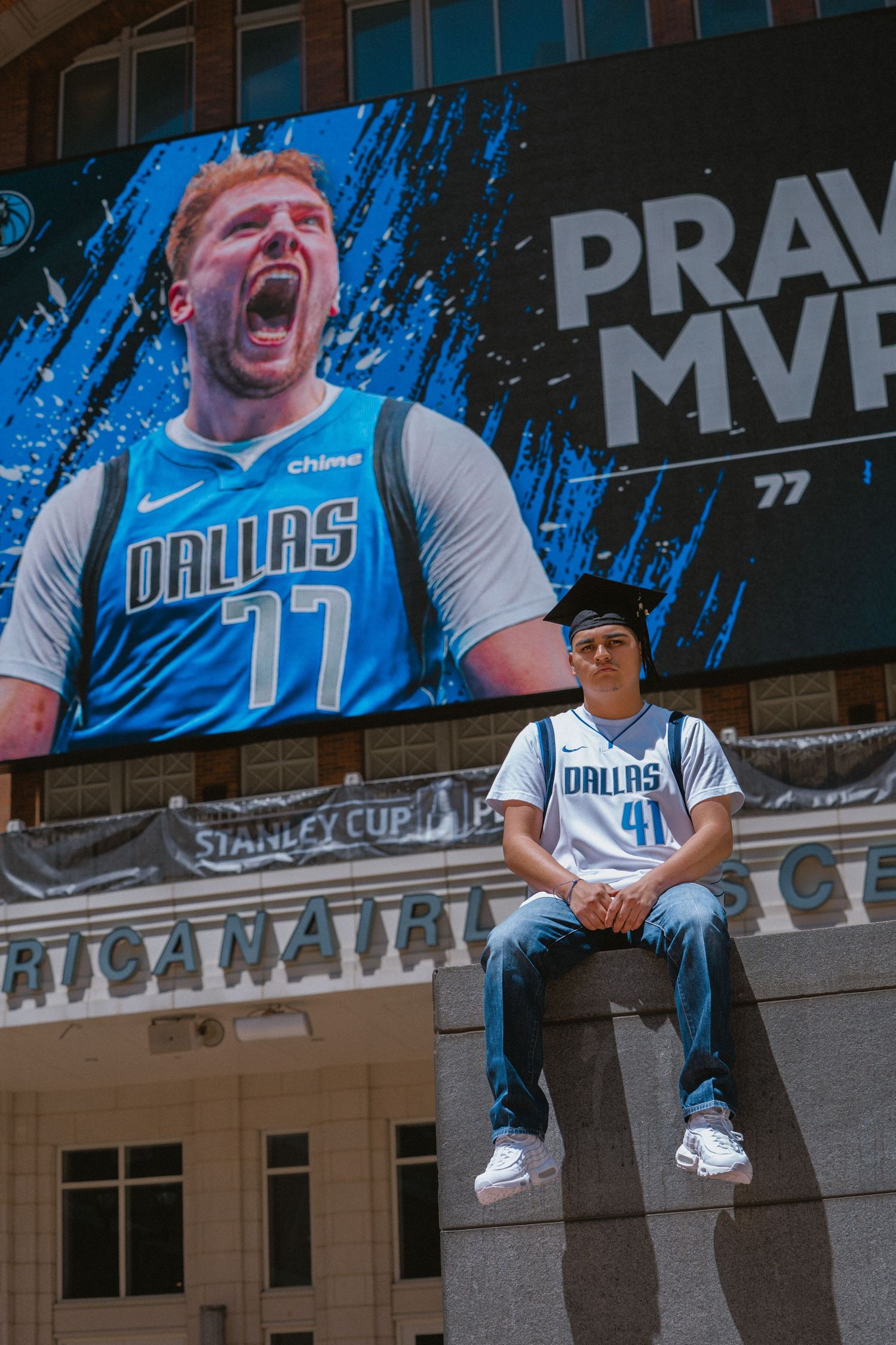 Dallas Mavericks fan in jersey #41 sits outside American Airlines Center beneath Luka Doncic MVP billboard.