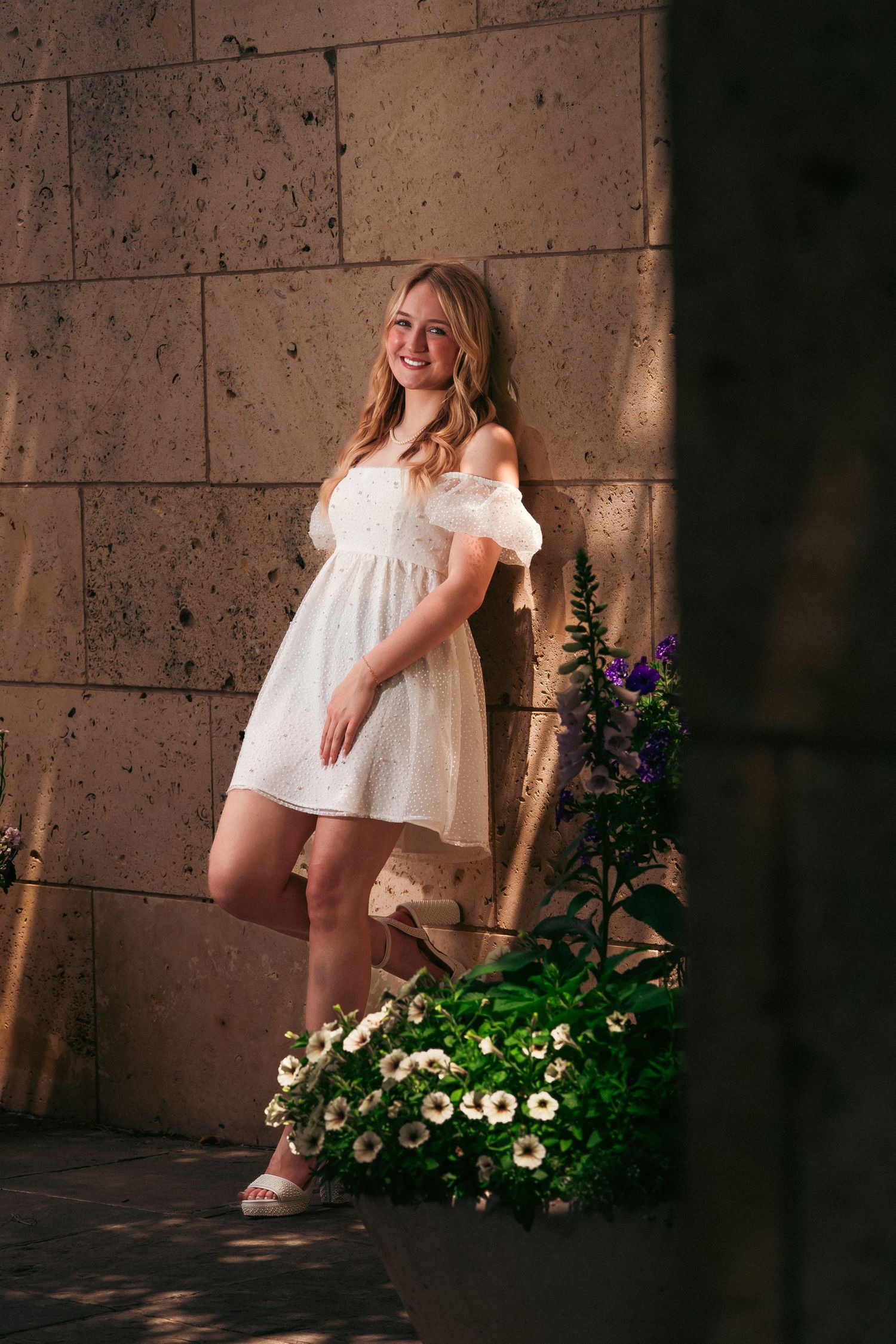 A series of four artistic maternity photos showing a woman in a white dress posing next to flowers against a concrete wall.