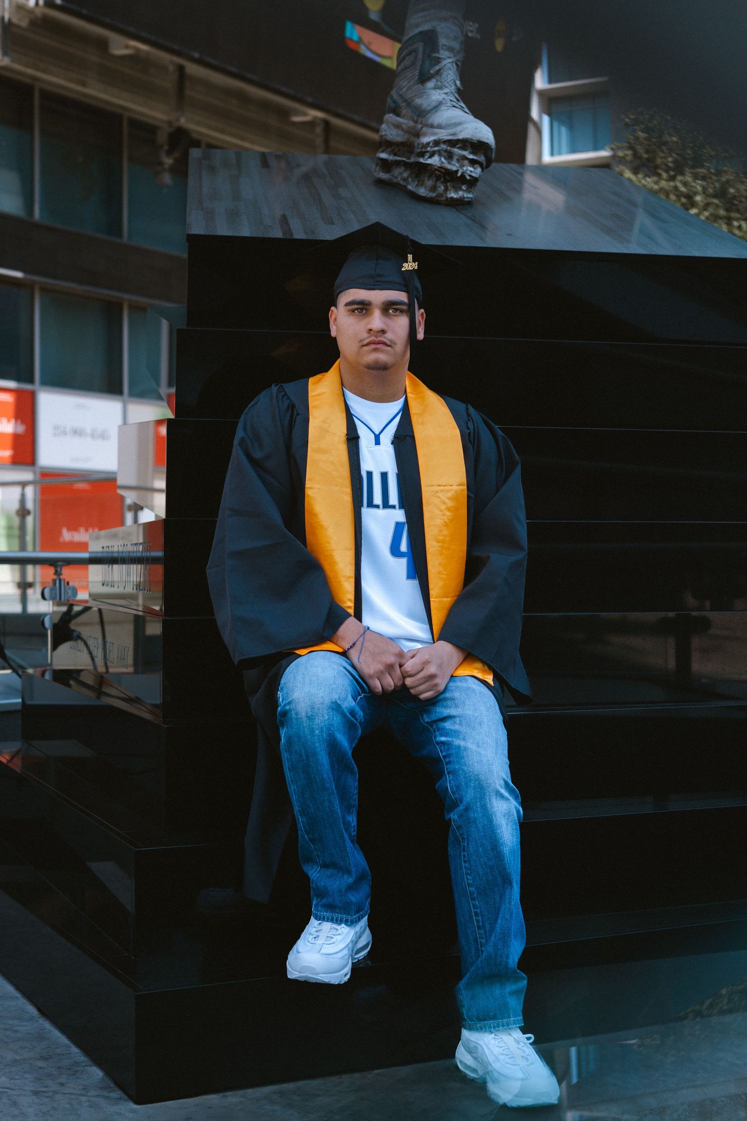 Graduate in cap, gown, and gold stole sits on dark monument steps wearing Dallas Mavericks jersey and jeans.