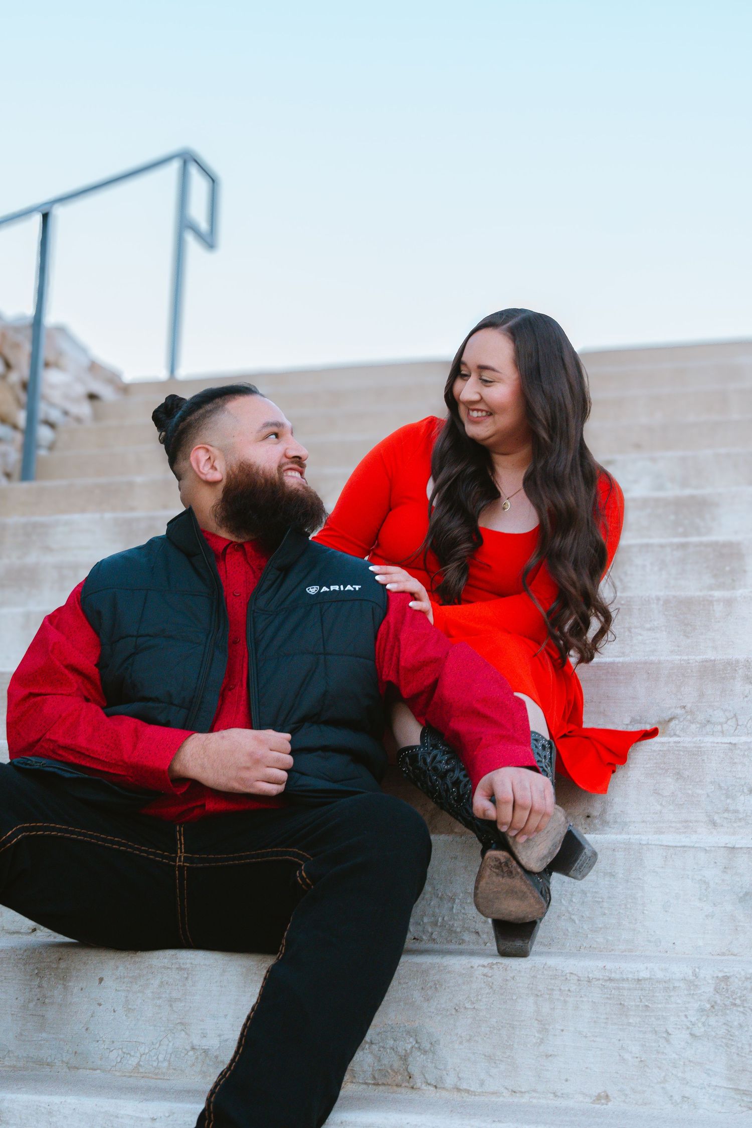 Couple sitting on outdoor steps smiling at each other, both wearing red outfits, man in black vest, woman in red dress.