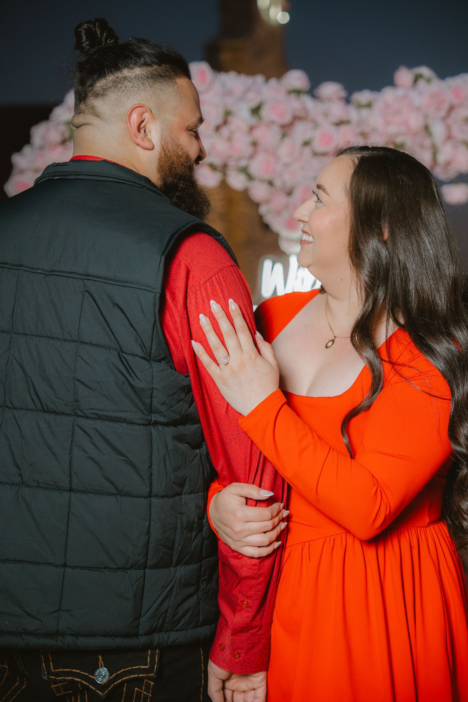 Couple in matching red and black outfits share a romantic moment during a marriage proposal with pink roses backdrop.