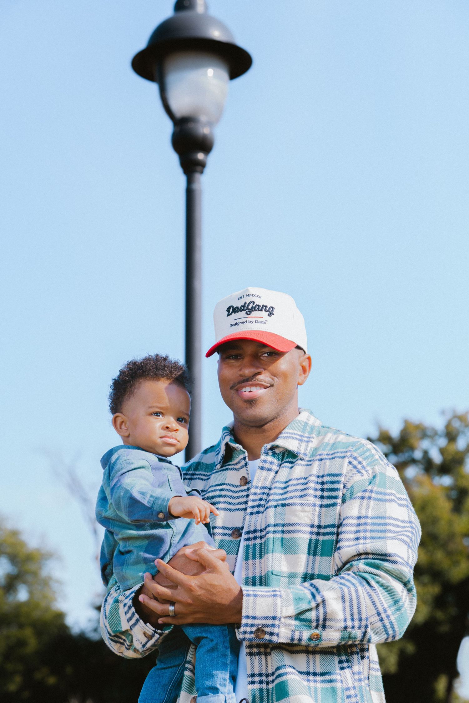 Proud Black father holding baby outdoors wearing plaid shirt and red DadGang cap under blue sky.