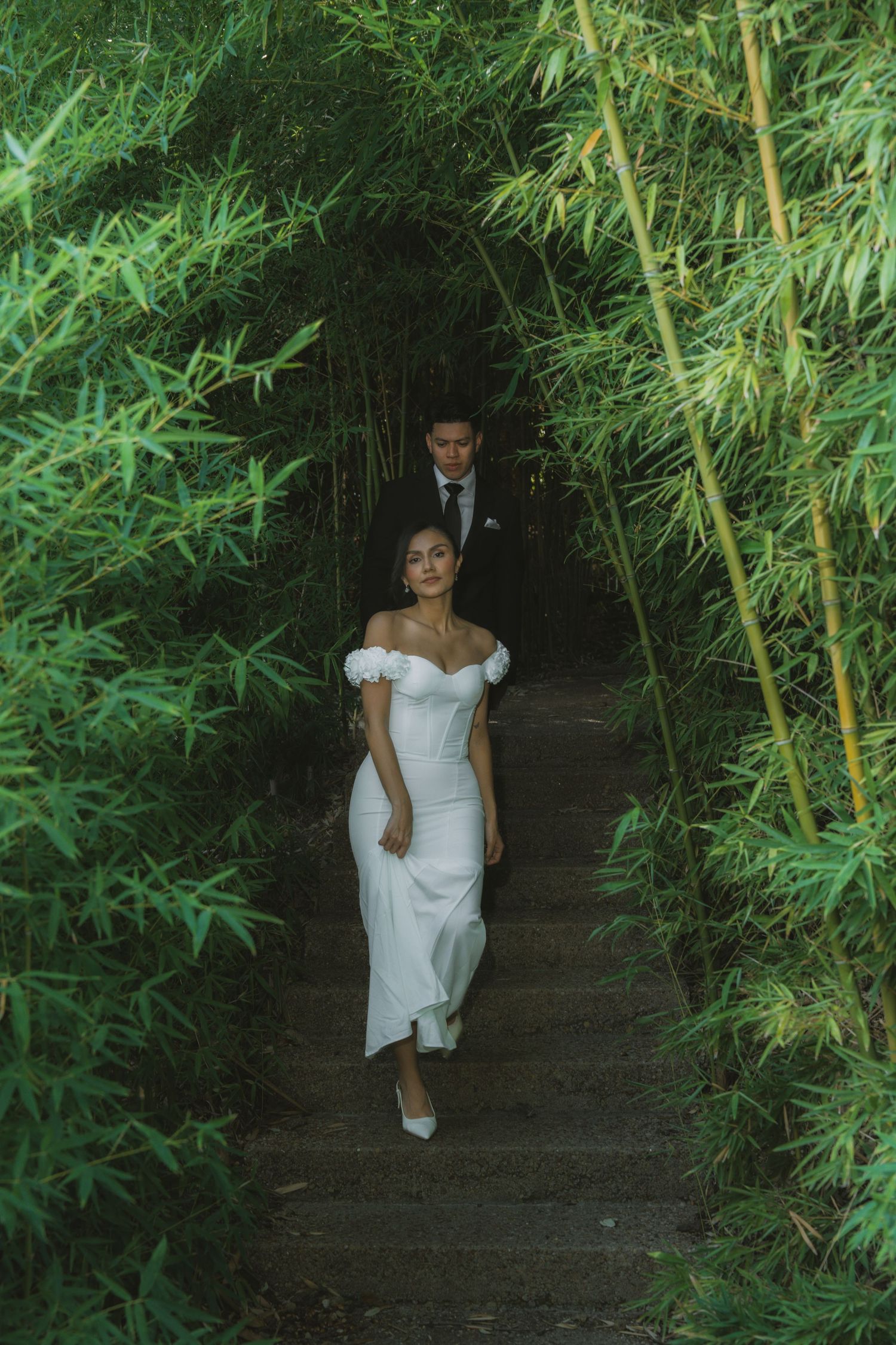 A couple descends stone steps surrounded by lush bamboo in wedding attire.