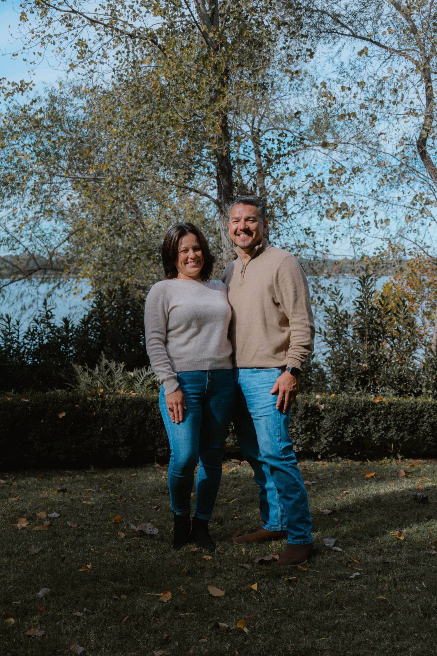 A couple in casual fall attire stands together on a grassy hillside with autumn trees in the background.