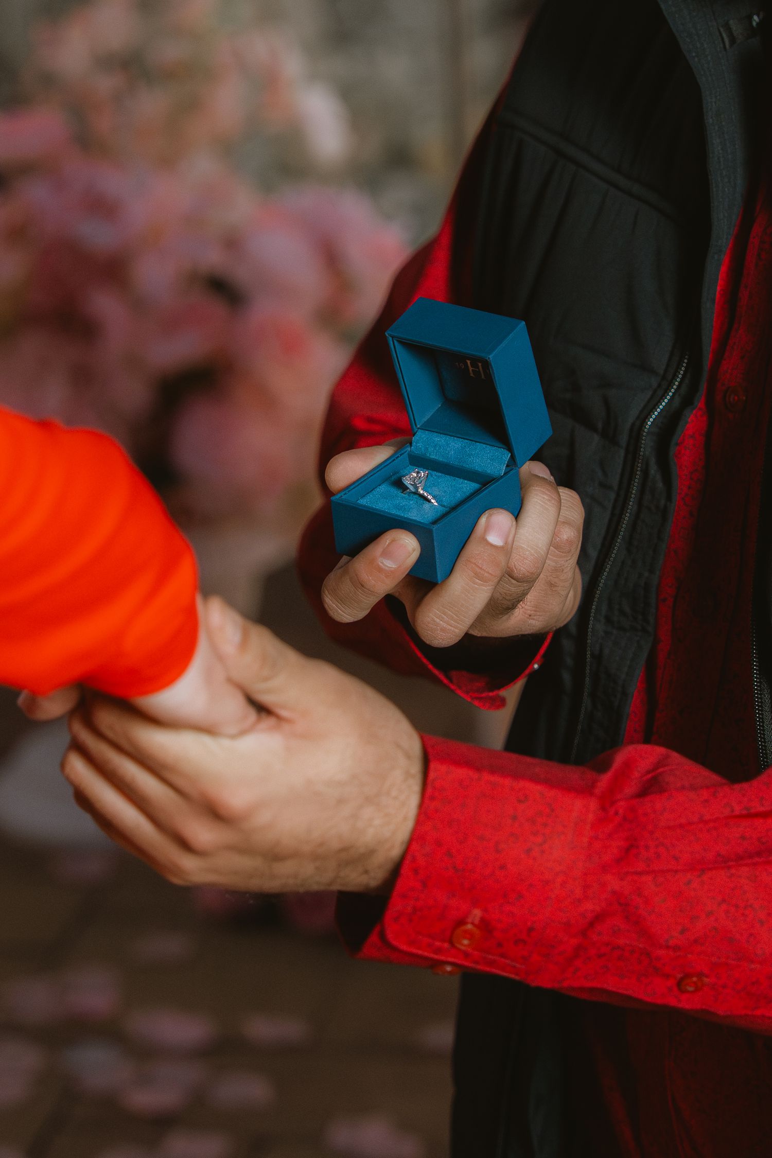 Man in red shirt holding blue velvet ring box during romantic marriage proposal outdoors.