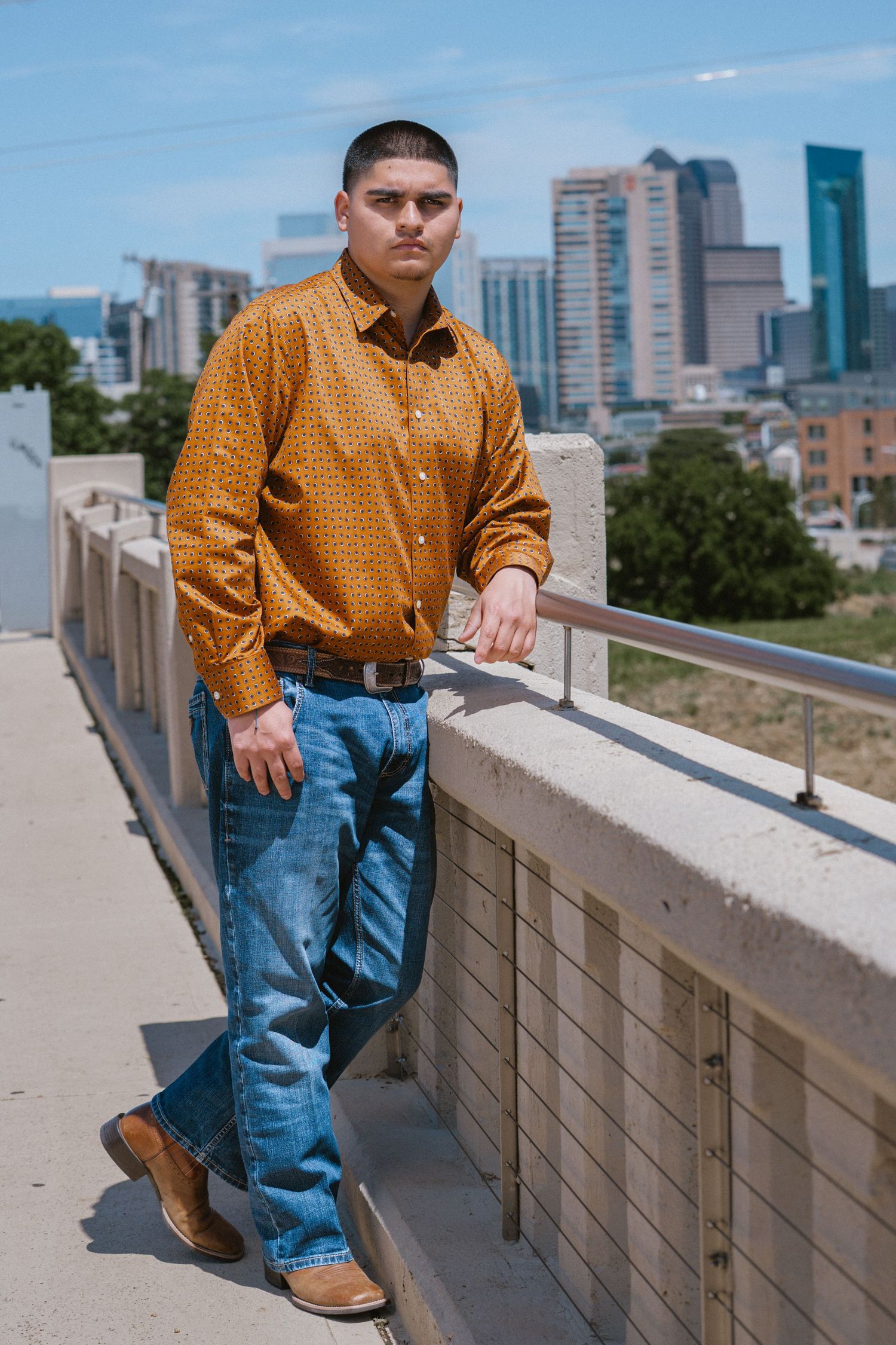 Young man in gold patterned shirt and jeans leaning on bridge railing with city skyline backdrop.