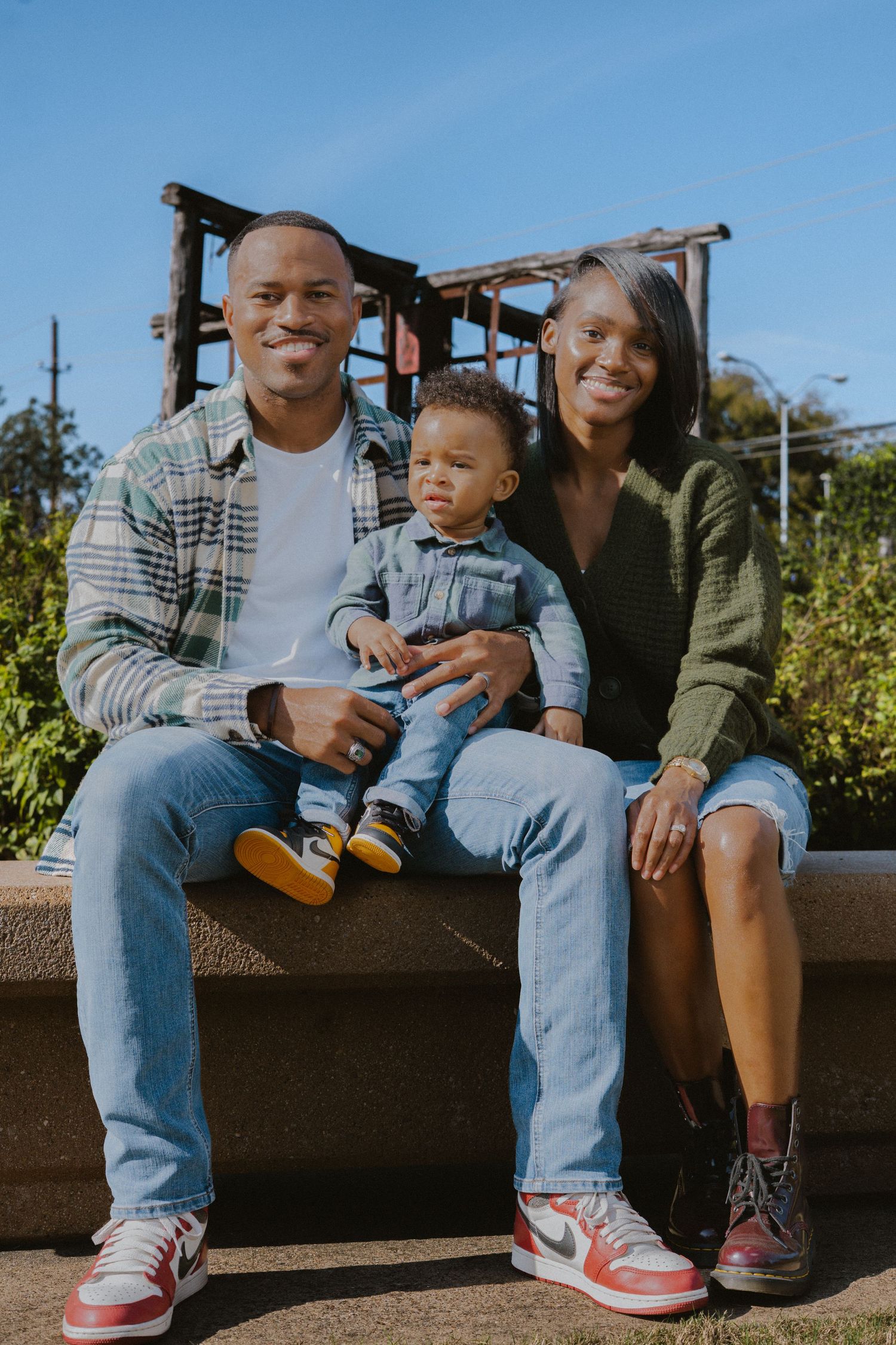 Happy Black family with baby sitting outdoors on sunny day, parents smiling in casual denim and plaid outfits.