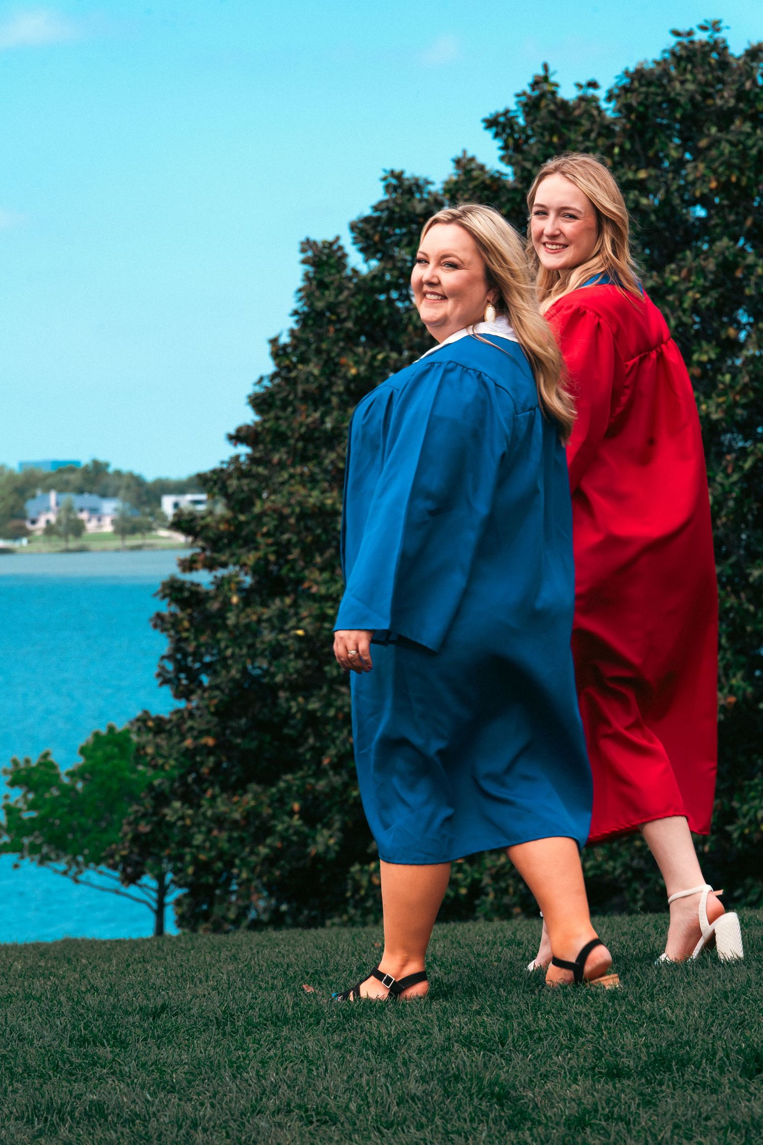 Two people in blue and red outfits share a playful moment on a grassy hill overlooking a scenic waterfront view.